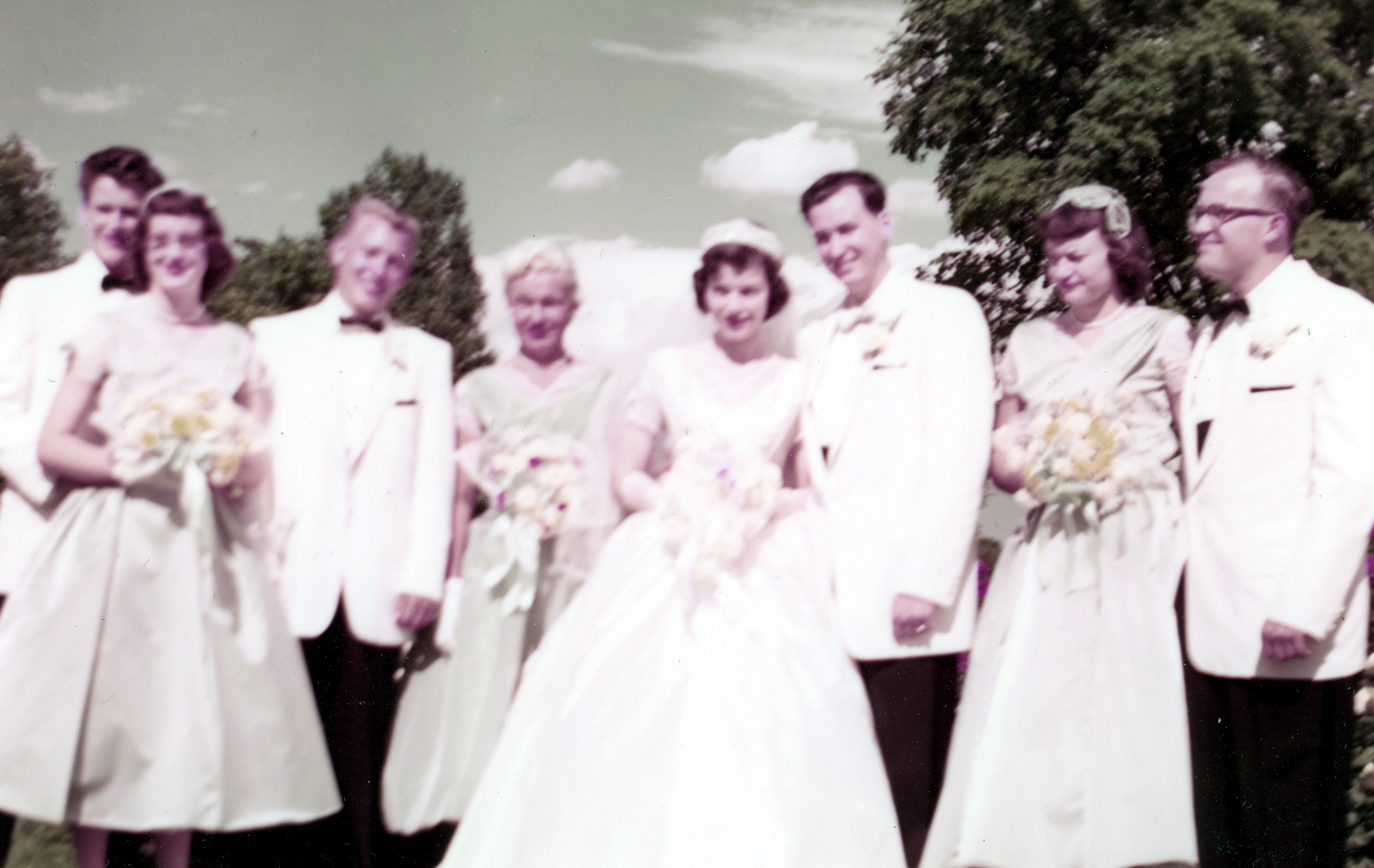 Wedding party outdoors: bridesmaids in white gowns holding bouquets and groomsmen in white jackets posing together on a sunny day.