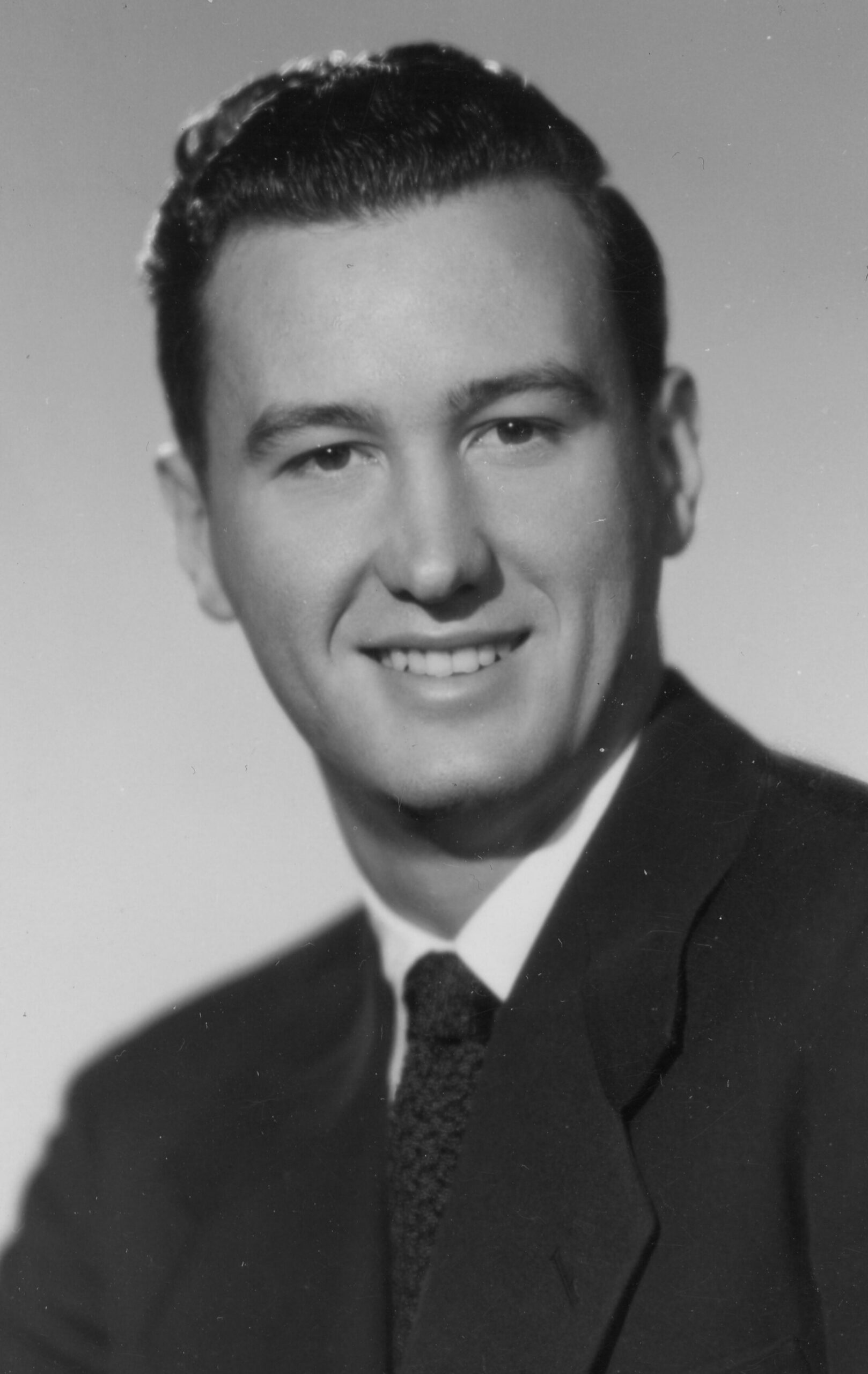Young man wearing a dark suit and tie, smiling in a studio portrait.