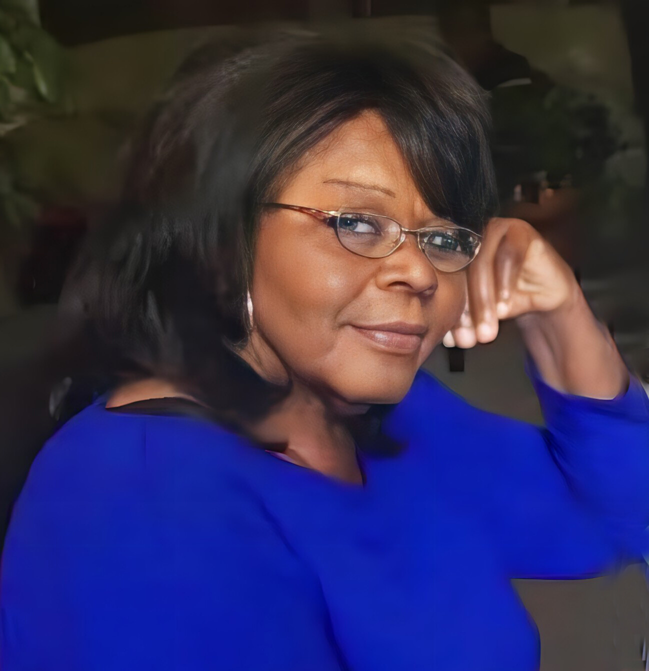 Portrait of a smiling woman wearing glasses and a royal blue top, resting her head on her hand.