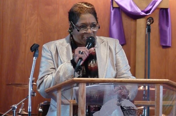 Woman in a beige blazer speaks into a handheld microphone at a wooden church pulpit with purple drapes in the background.