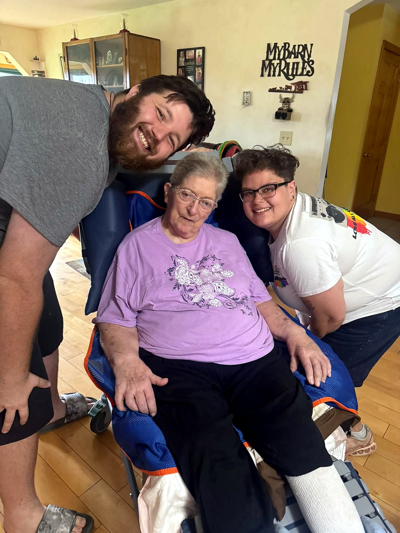 Three people smile at the camera around an elderly woman in a purple shirt seated in a wheelchair indoors, with two helpers on either side—friendly family-like moment to capture.