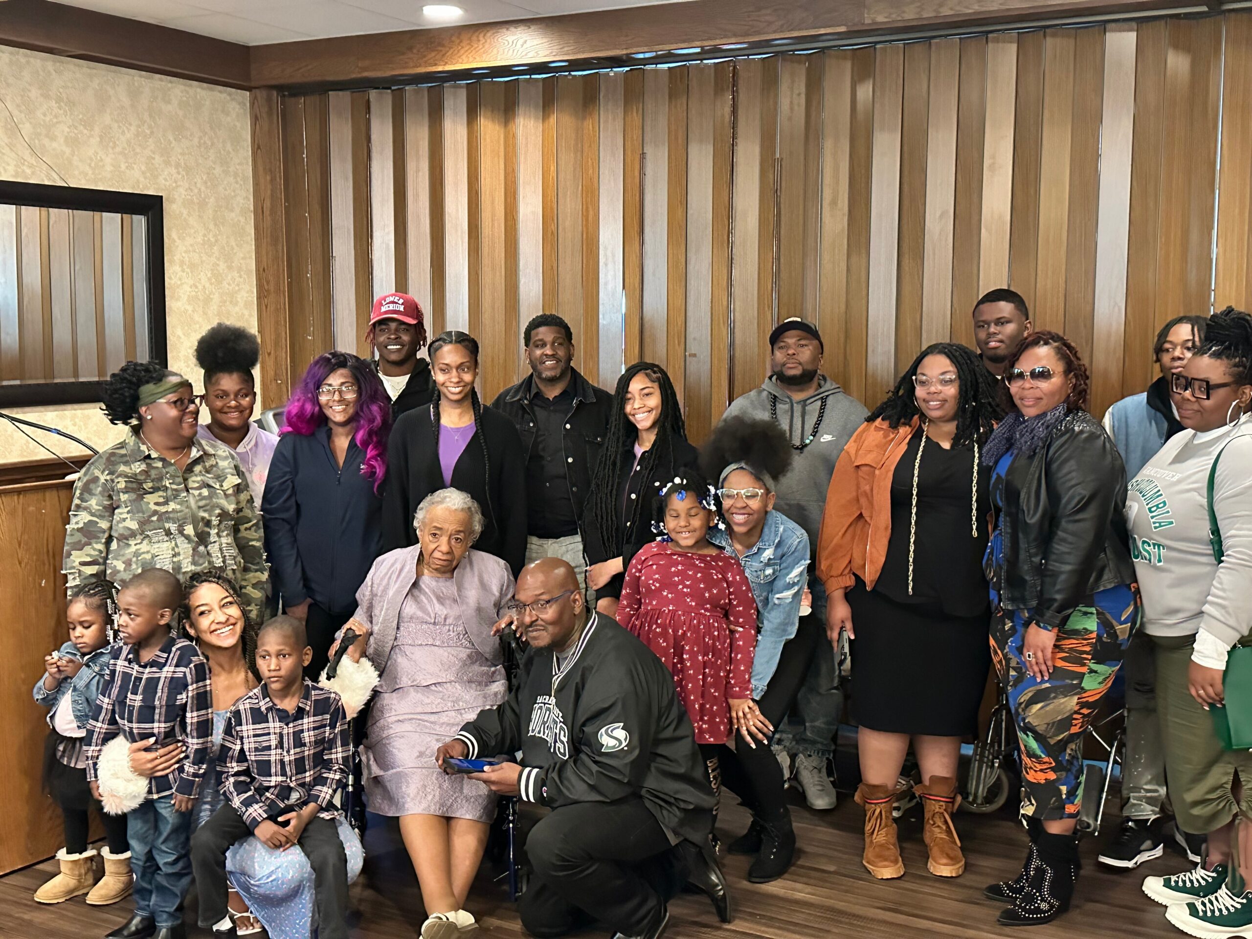 Group portrait of a diverse crowd of adults and children posing together indoors in front of wood-paneled walls. Many are smiling at the camera.