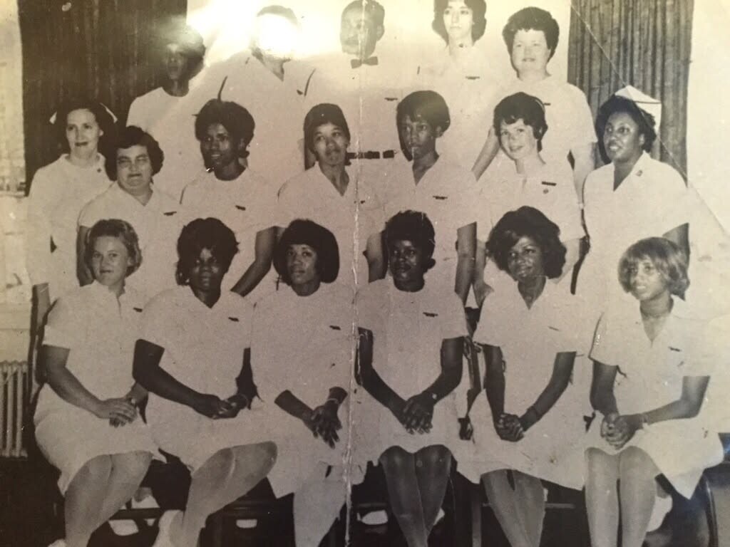 Group of women in white nurses' uniforms posing for a class photo in a hospital-like room, seated and standing in rows.