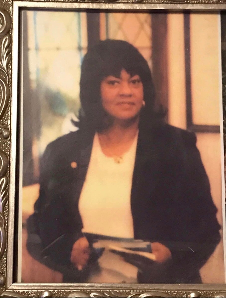Portrait of a woman with dark hair in a black blazer and white blouse, holding a booklet, seated indoors in a framed photograph state.