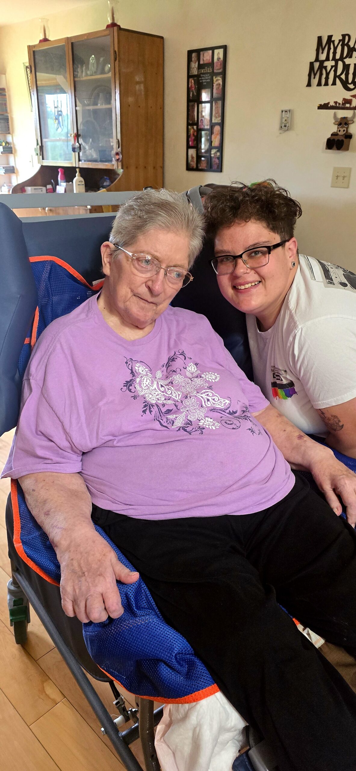 Older woman with gray hair and glasses sits in a blue wheelchair, wearing a purple shirt, smiling next to a younger caregiver in a white shirt at home.