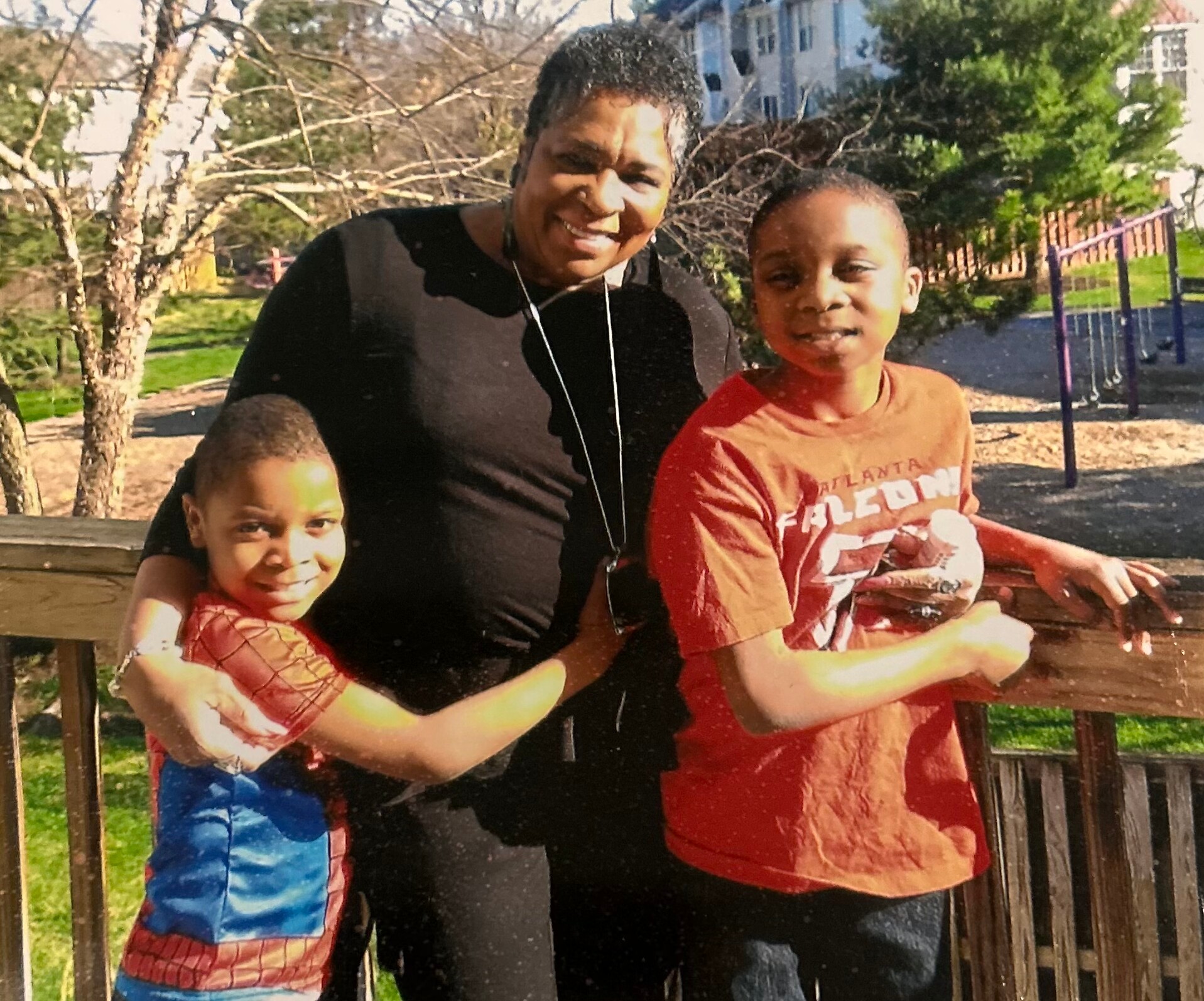 Smiling woman in a black top with two children outdoors, one in a Spider-Man outfit and the other in a red shirt, beside a wooden railing at a park.