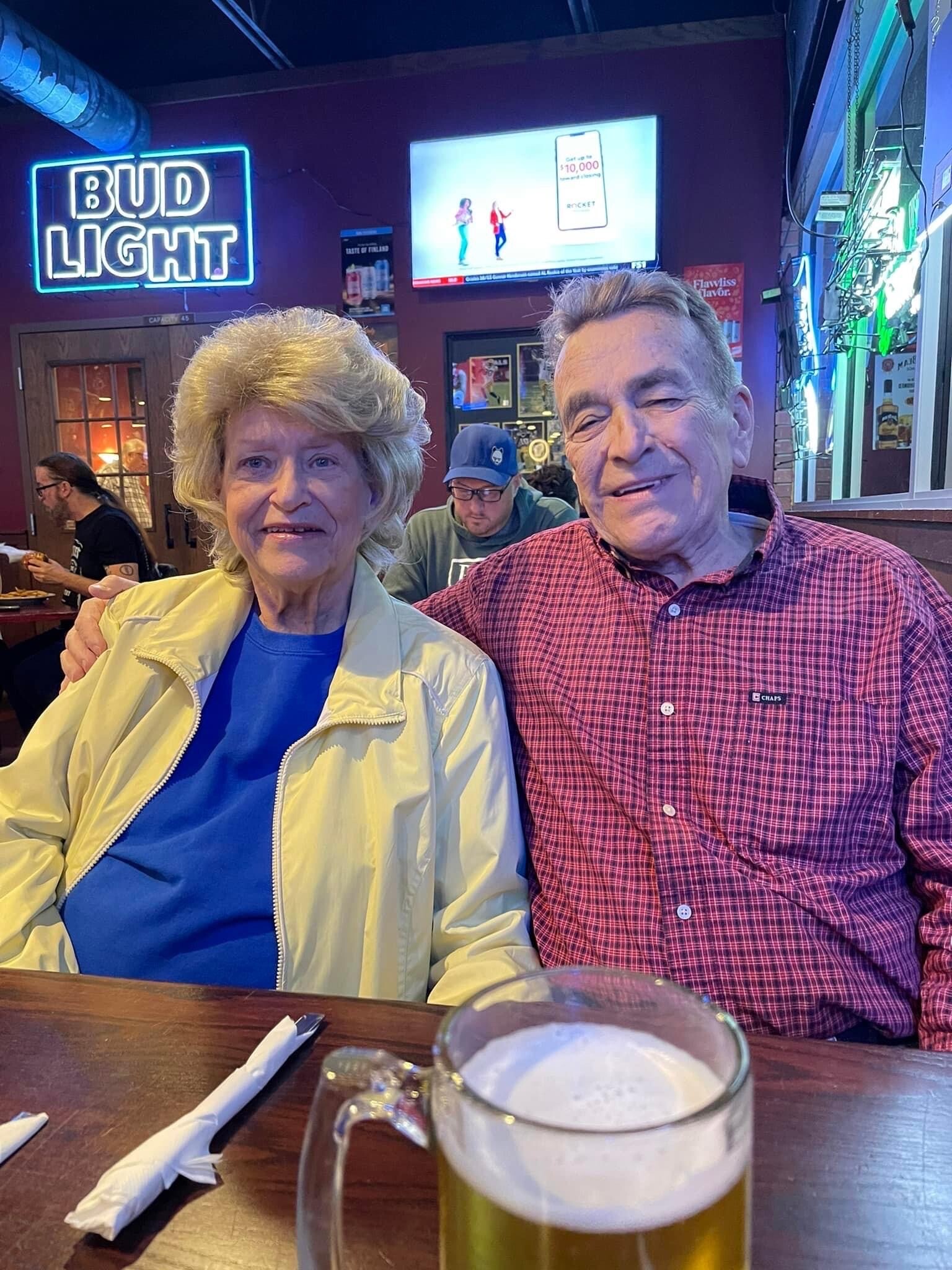 Two smiling seniors sit close together at a restaurant table, a glass of beer in the foreground, with a Bud Light neon sign behind them and a TV on the wall in the background.