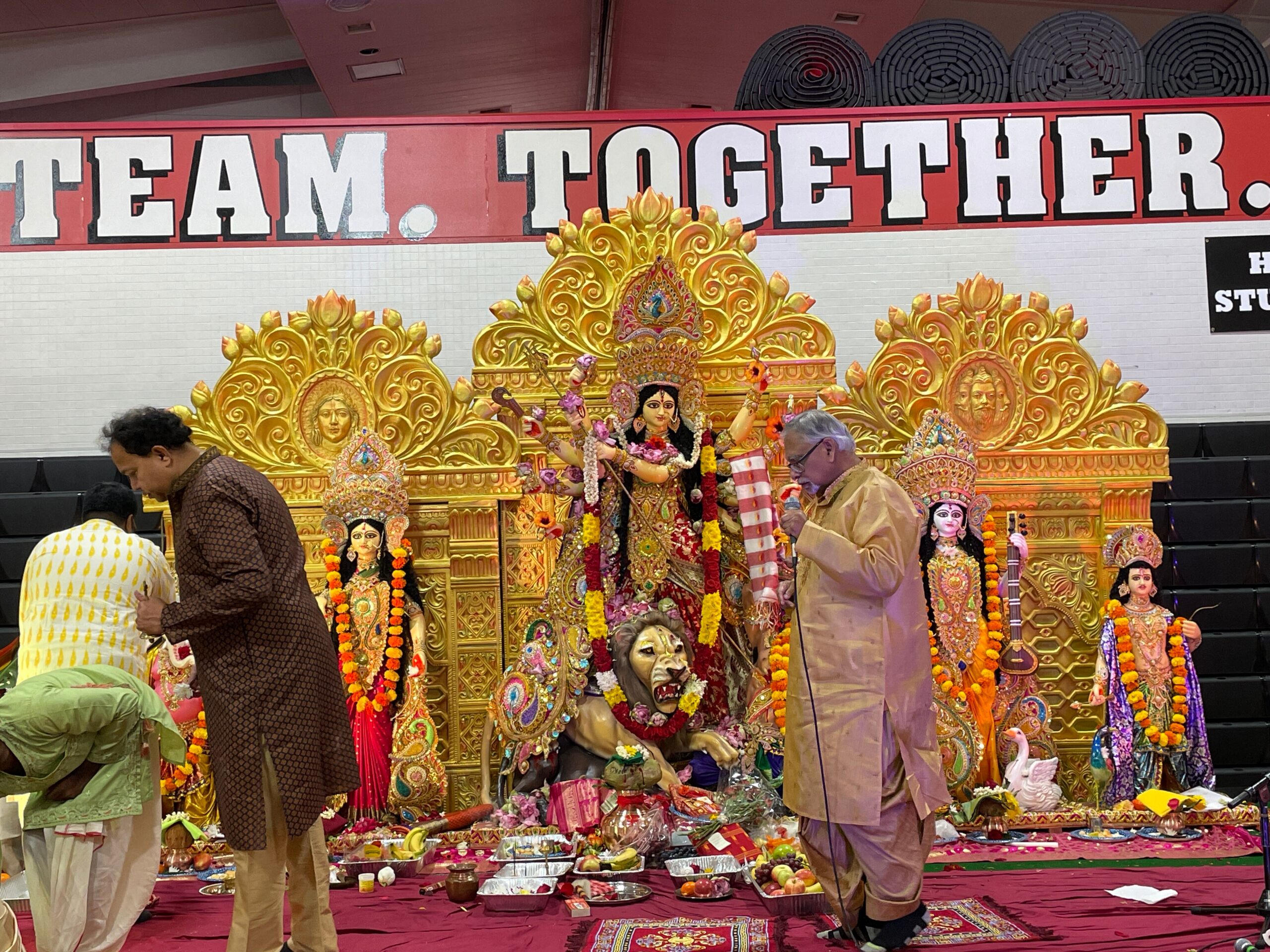 Durga Puja altar with ornate gold idols and floral garlands, people in traditional dress arranging offerings on a red carpet.