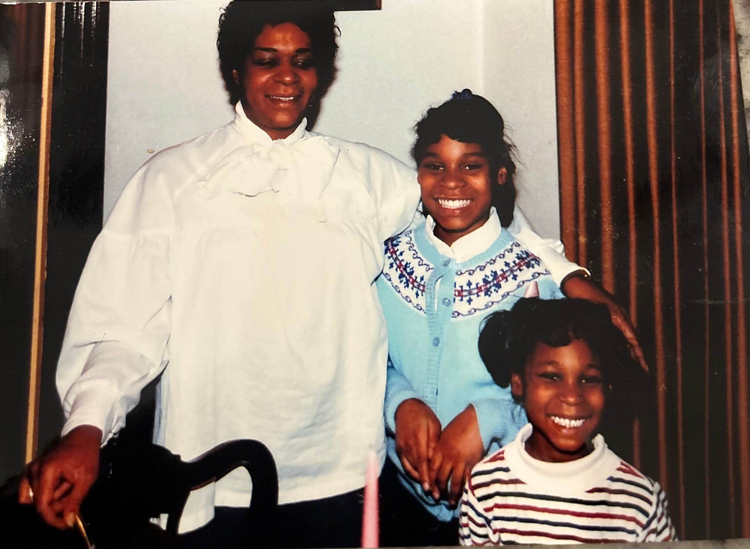 Smiling woman in a white blouse standing with two children, posing together for a family photo indoors.