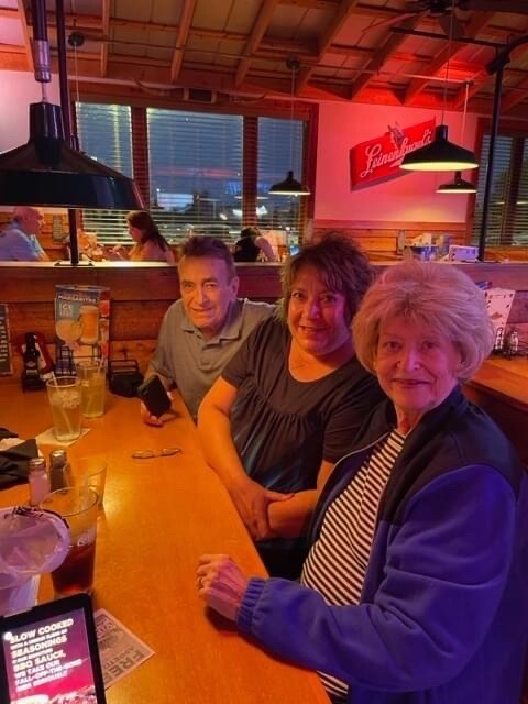Three adults sit at a wooden bar in a cozy restaurant, smiling at the camera with drinks on the counter.