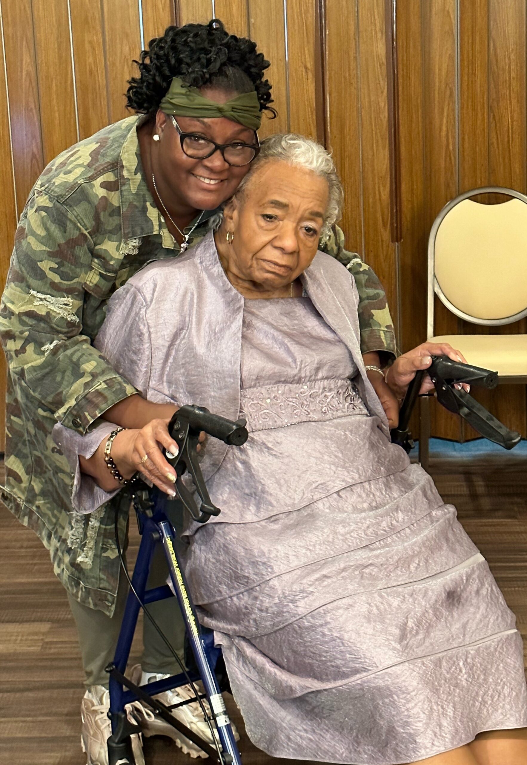 Caregiver in camo jacket leaning over an elderly woman in a light gray dress seated in a wheelchair indoors.