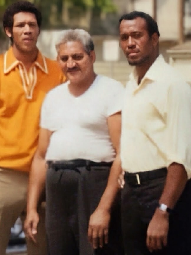 Three men posing together outdoors, one in white t-shirt center, flanked by a man in orange shirt left and a man in light yellow shirt right.