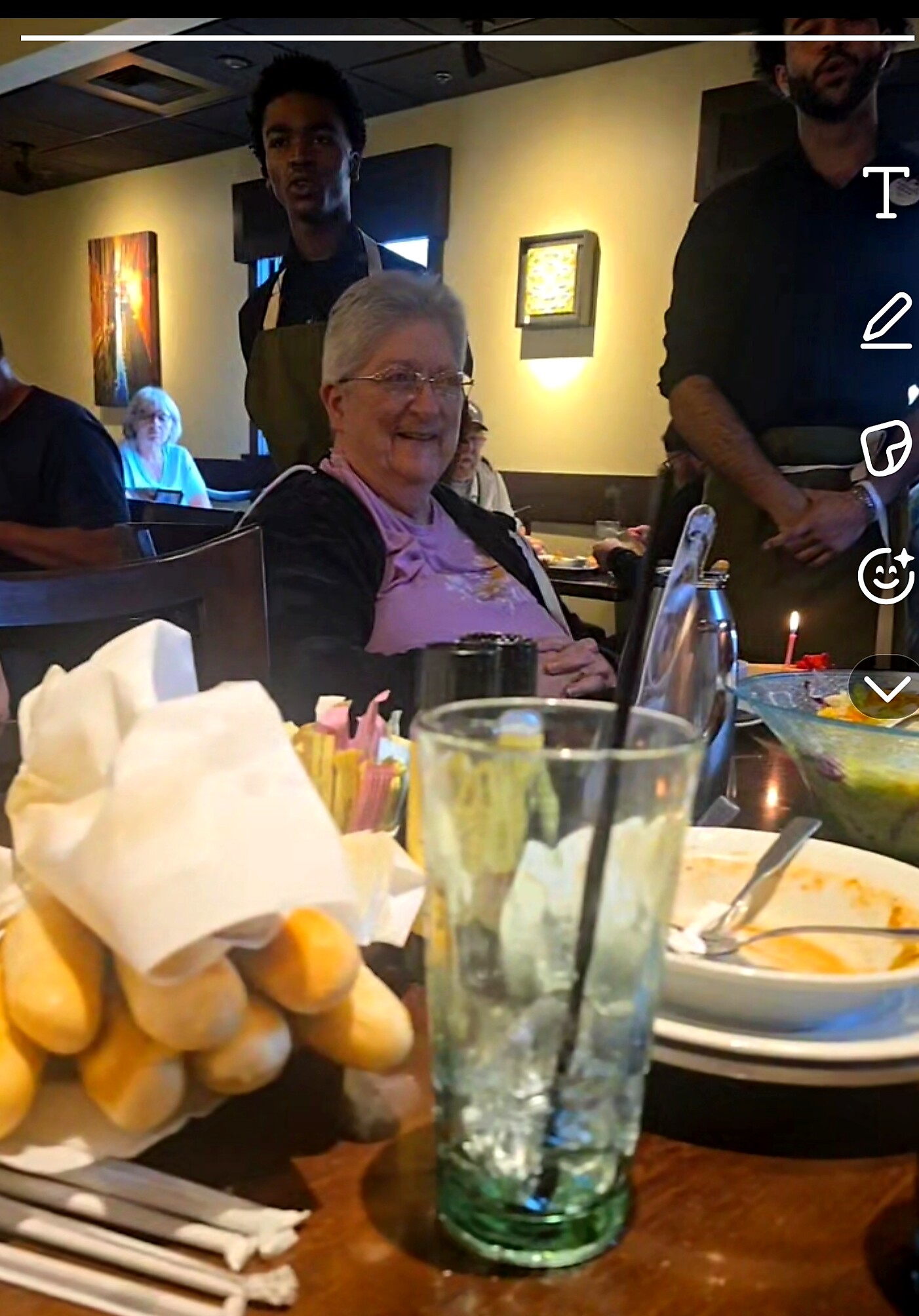 Older woman smiling at a restaurant table with bread basket, cups, and plates in front of her; others dine in the background.