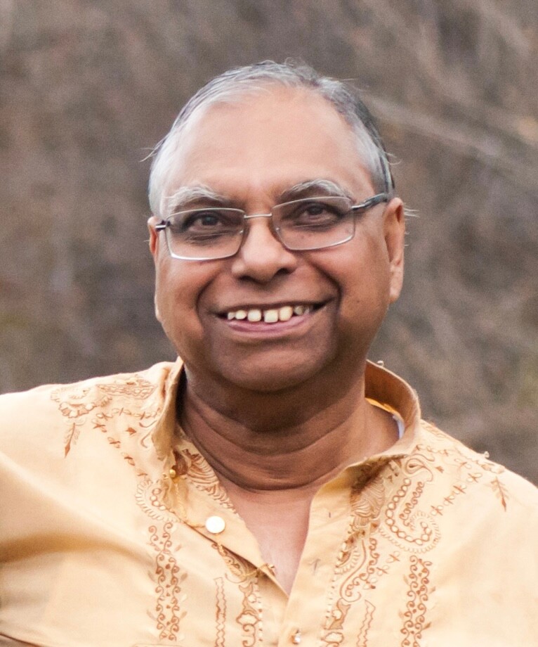 Smiling older man with gray hair and glasses wearing a beige patterned shirt, outdoors.