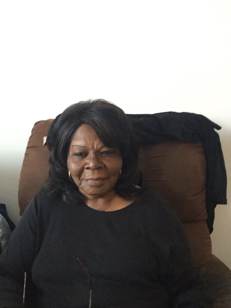 Older woman with dark hair and hoop earrings sits in a brown recliner, wearing a black top indoors, glasses resting on her chest.