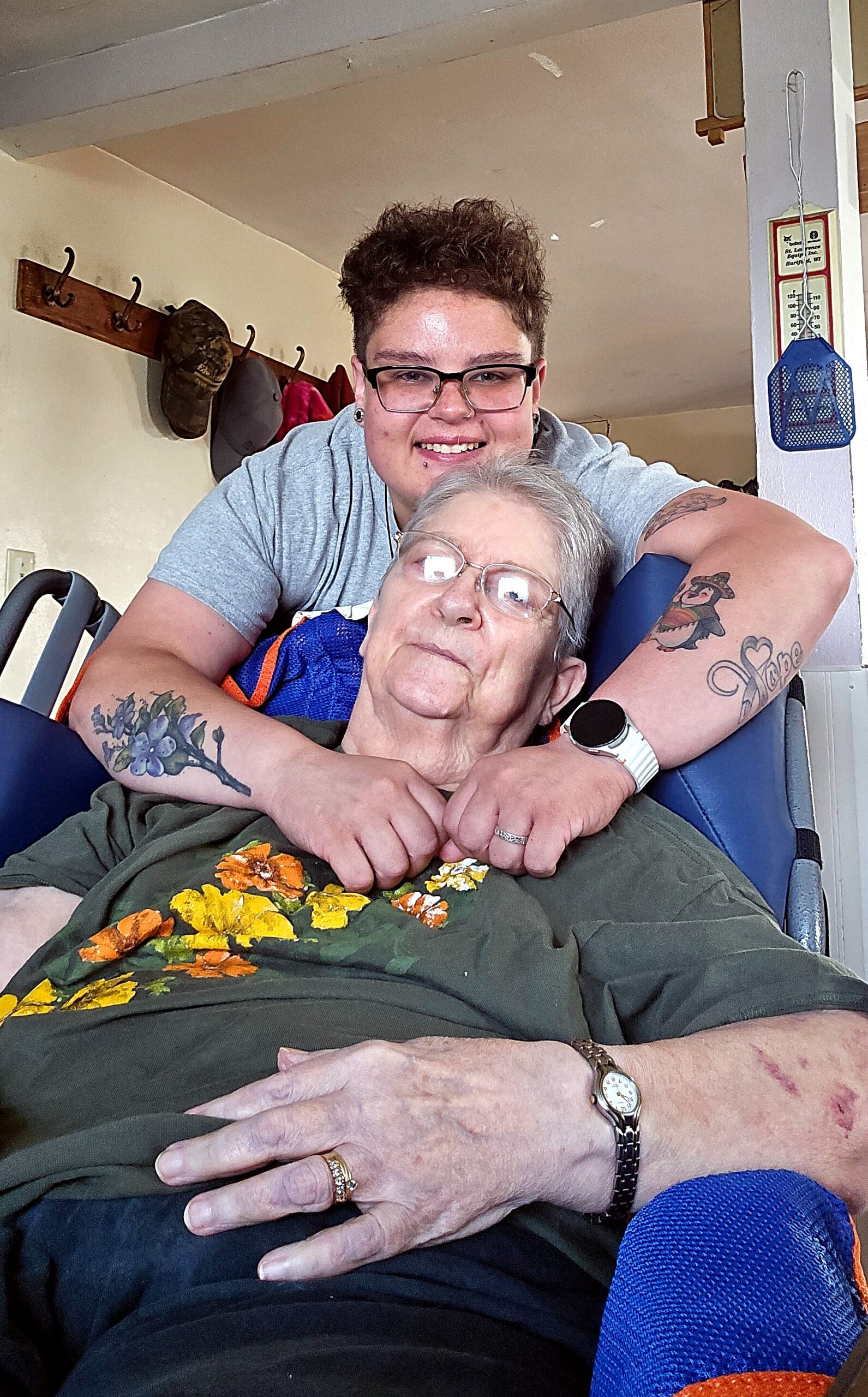 Young person with short hair and glasses hugs an elderly woman seated in a blue chair, both smiling in a home setting.