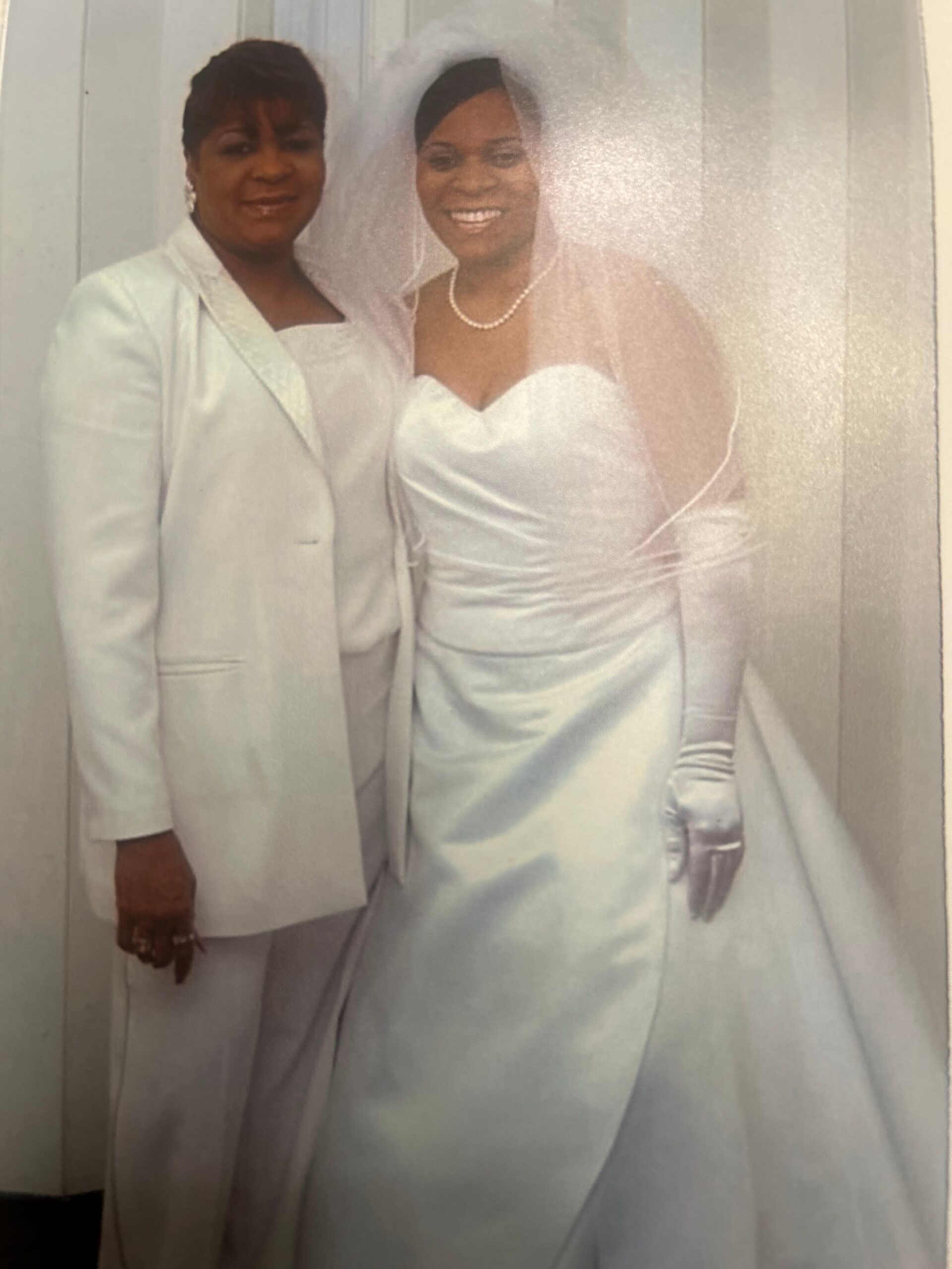 Two women in formal attire pose together: a bride in a white strapless gown with veil and pearl necklace beside a guest in a pale suit.
