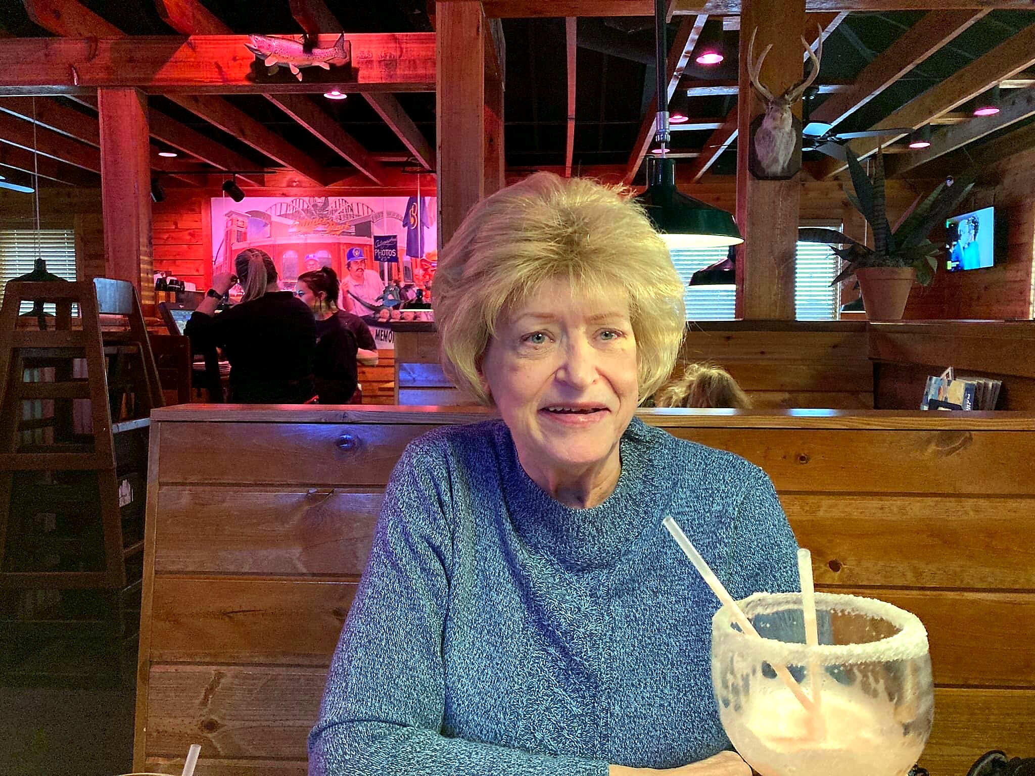 Older woman in a blue sweater sits at a wooden booth in a rustic restaurant, with a pale drink and striped straw in front of her.