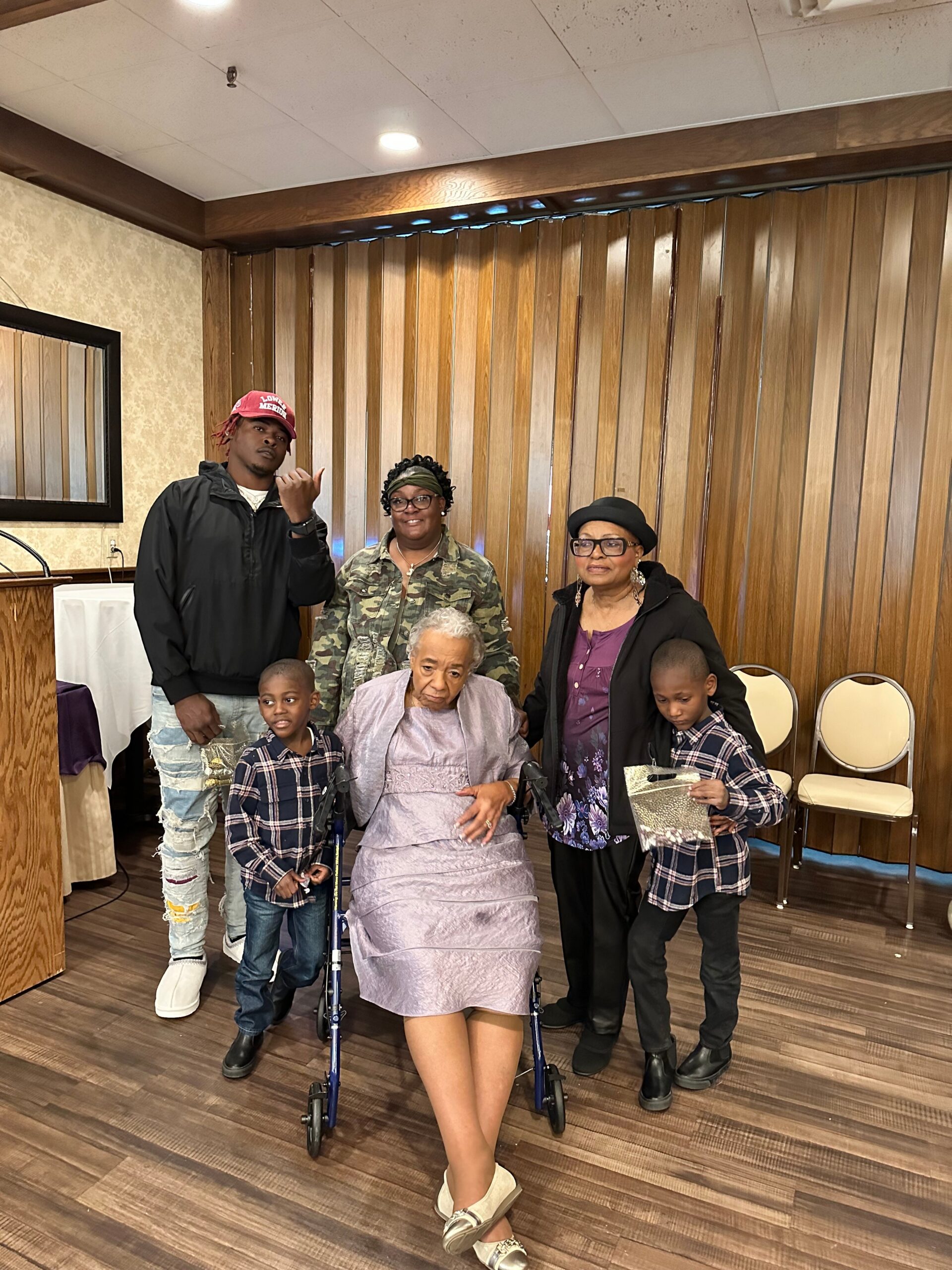 Family group indoors posing for a photo around an elderly woman in a lavender dress and wheelchair, with adults and children nearby.