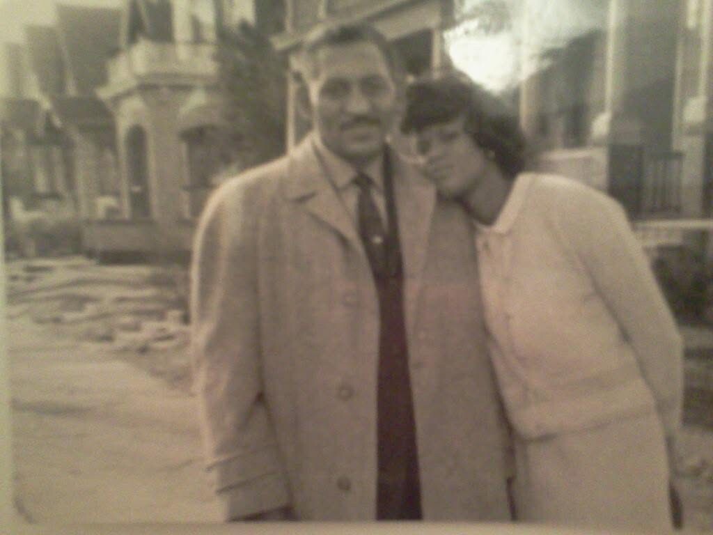A man in a long coat and tie stands with a woman leaning her head on his shoulder on a sidewalk, posing for a photo on a quiet street.