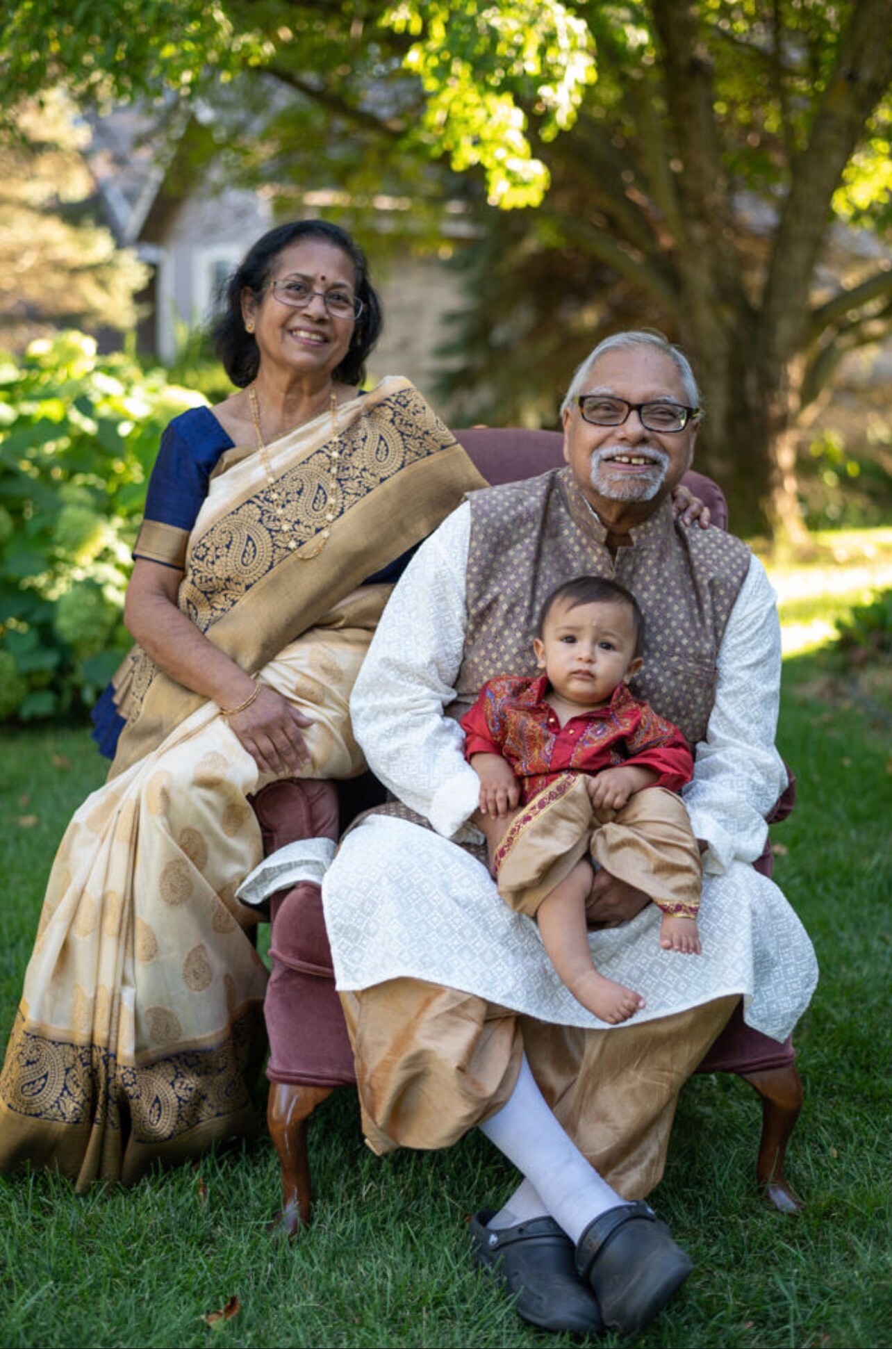Grandparents in traditional Indian clothing sit outdoors with their baby grandson, smiling in a sunlit garden.
