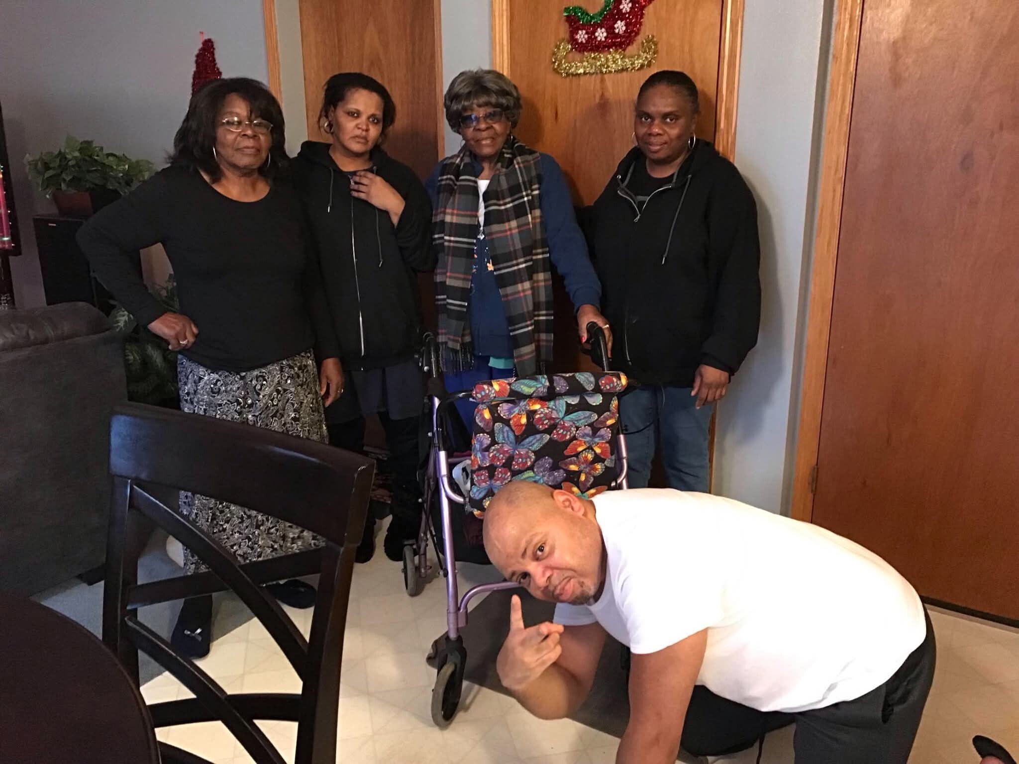 Four adults pose in a living room; a man kneels in front using a walker, holiday decor on the door behind them.
