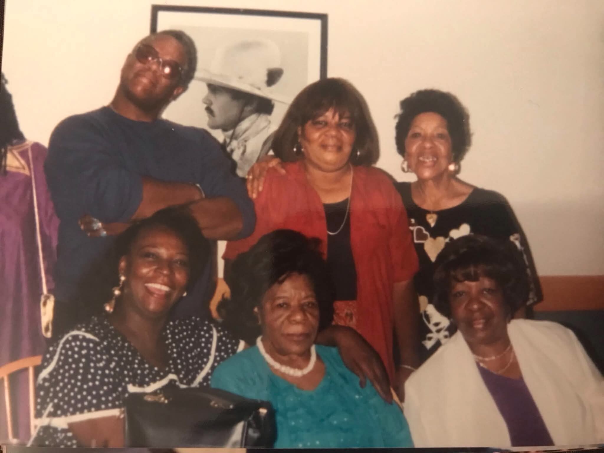 Group of six adults posing indoors, smiling, dressed in colorful outfits with a framed picture on the wall behind them.