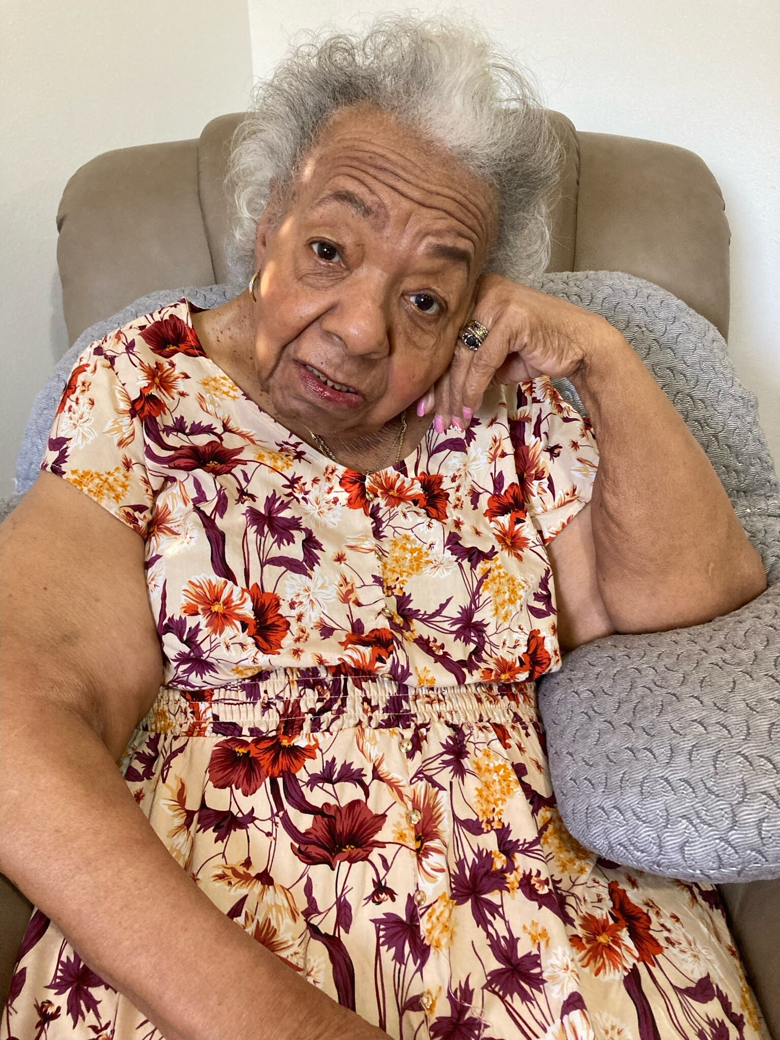 Older woman with gray hair rests her head on her hand while seated in a beige chair, wearing a colorful floral dress.