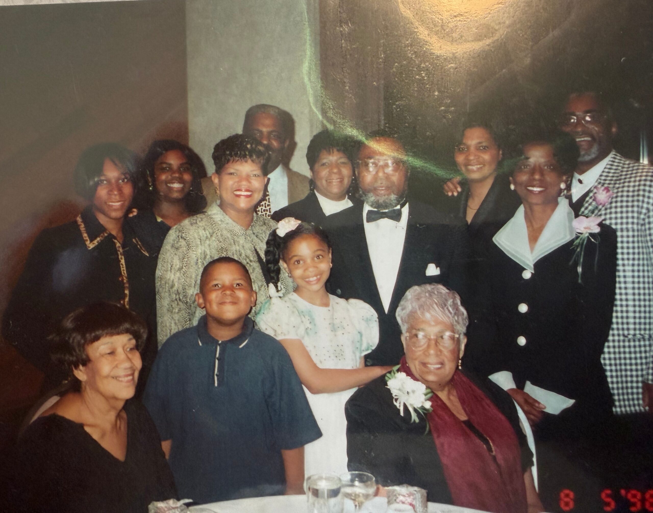 A large group of adults and children smiling for a group photo at a formal celebration, dressed in dresses and suits around a table barrier.