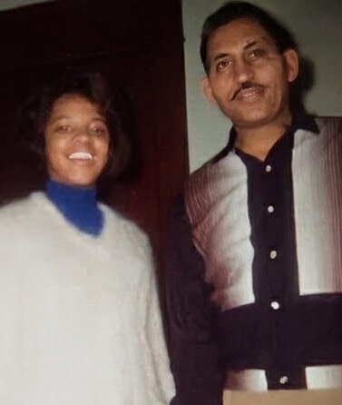 Two people standing side by side indoors, a smiling woman in a white sweater next to a man in a black and white striped shirt and dark pants.