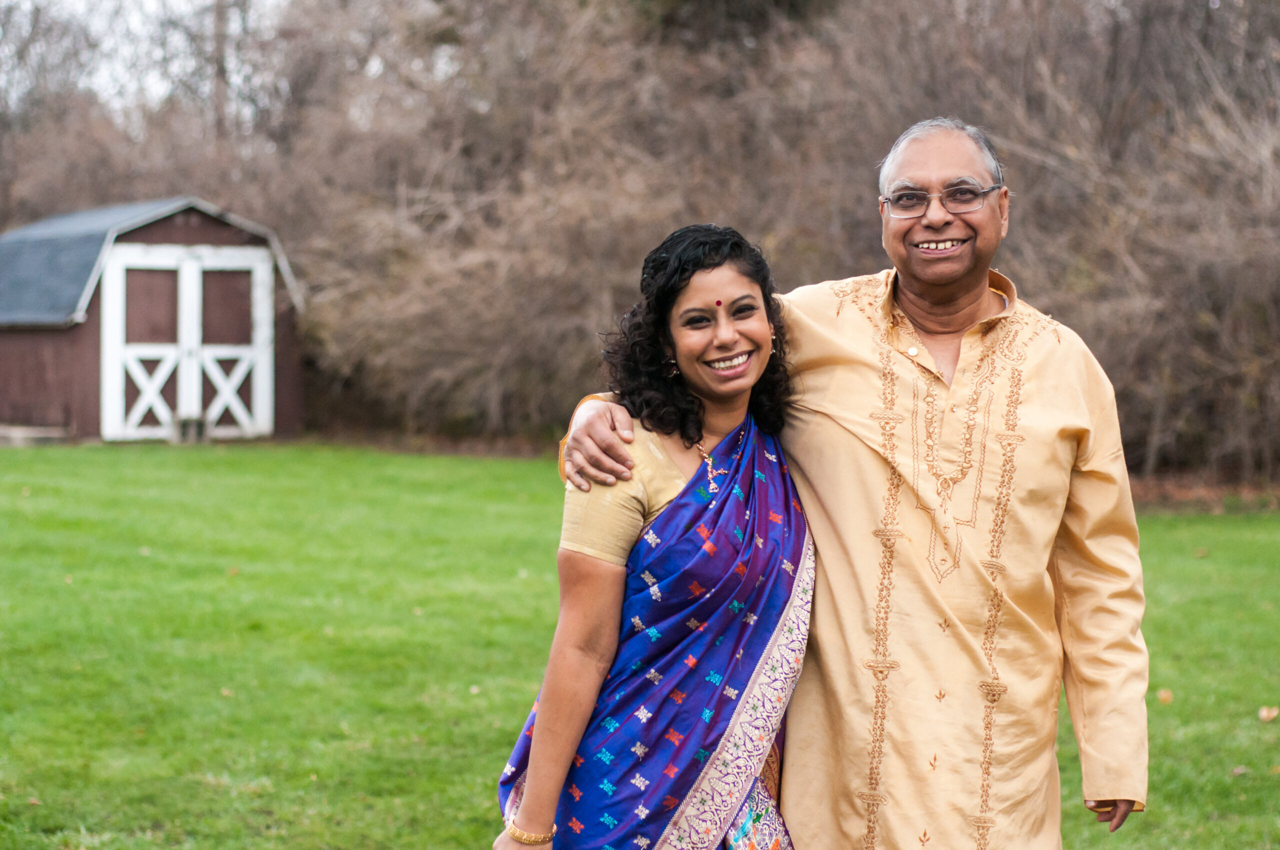 Smiling couple with arms around each other in a park; man in beige traditional shirt, woman in blue sari, a shed in the background.