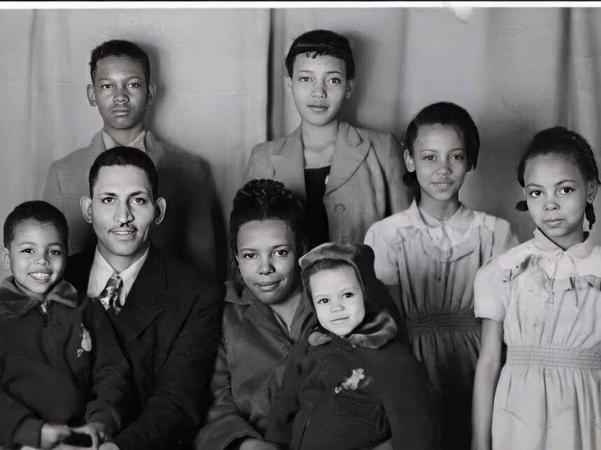 Group family portrait featuring three adults and five children seated and standing together indoors; everyone looks at the camera (black-and-white photo).”],