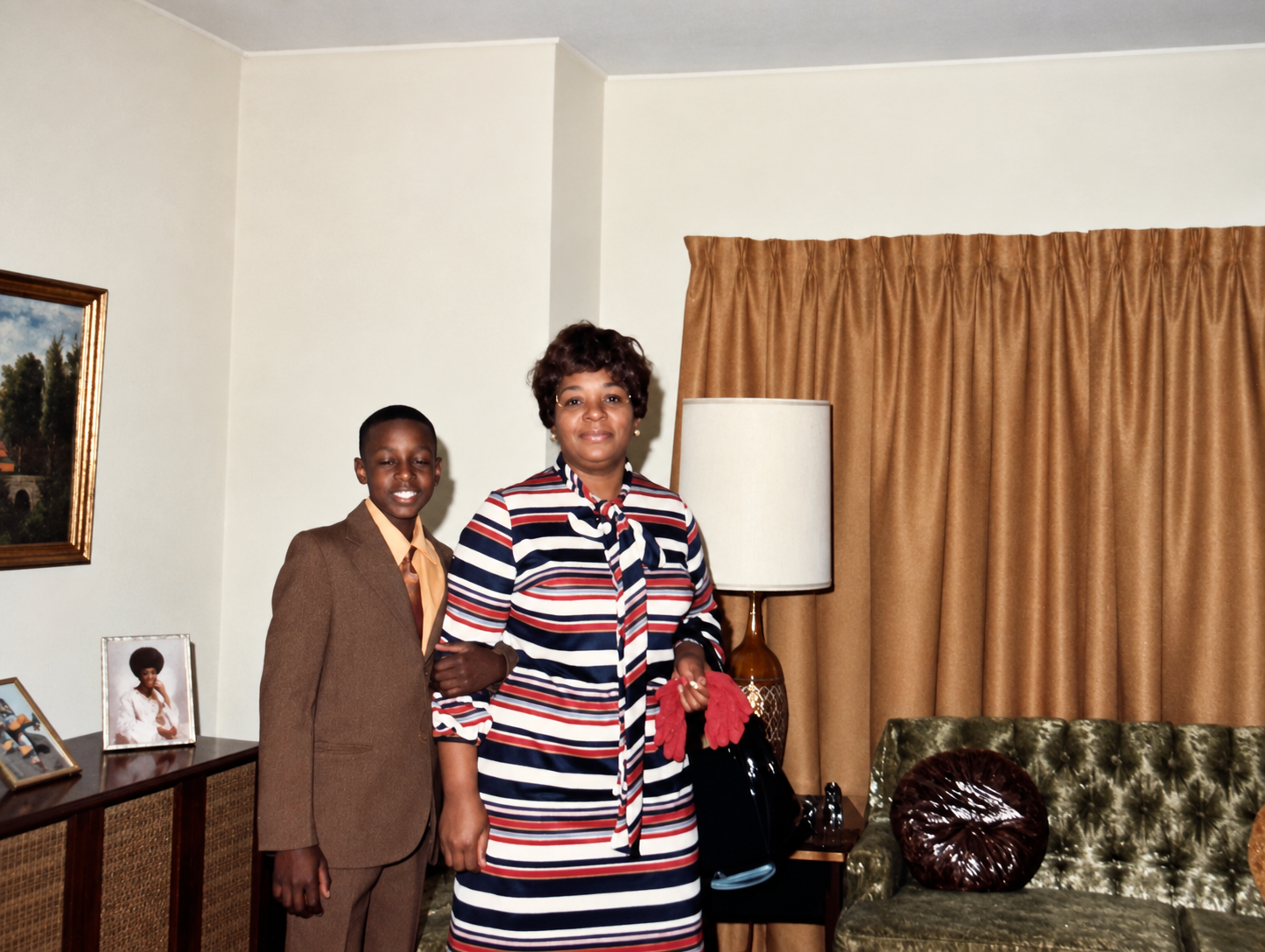 A woman and a boy pose together in a living room with brown curtains, a tufted green sofa, and framed photos on a sideboard behind them.