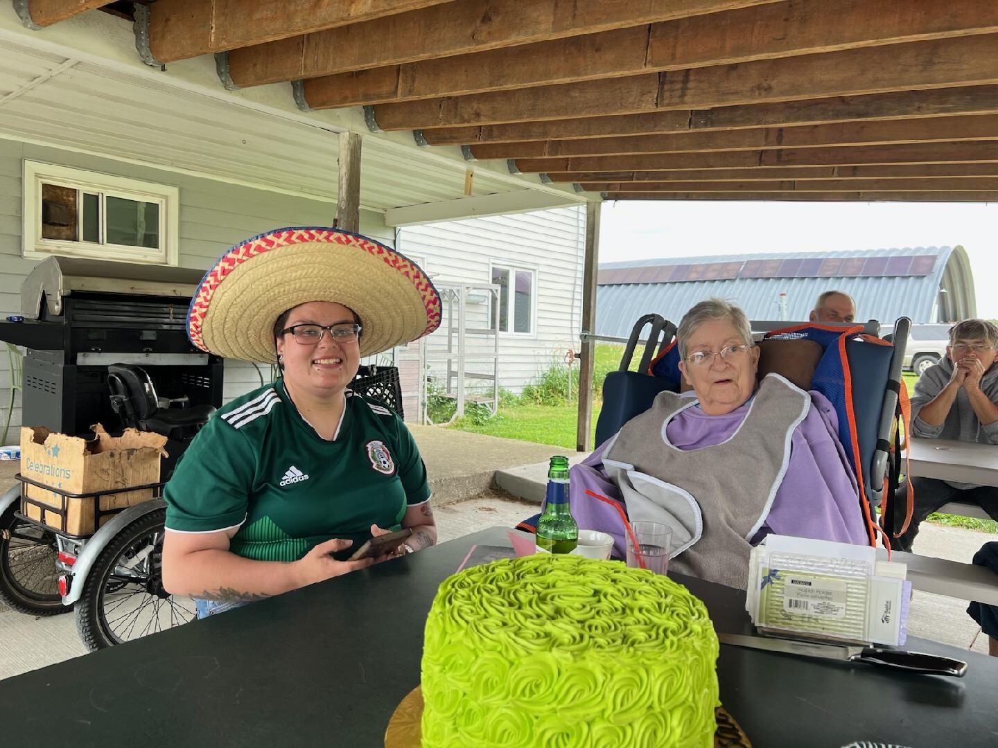 Woman wearing a large sombrero sits at a table with a bright green frosted cake in front; an elderly person in a wheelchair watches nearby under a covered outdoor area.