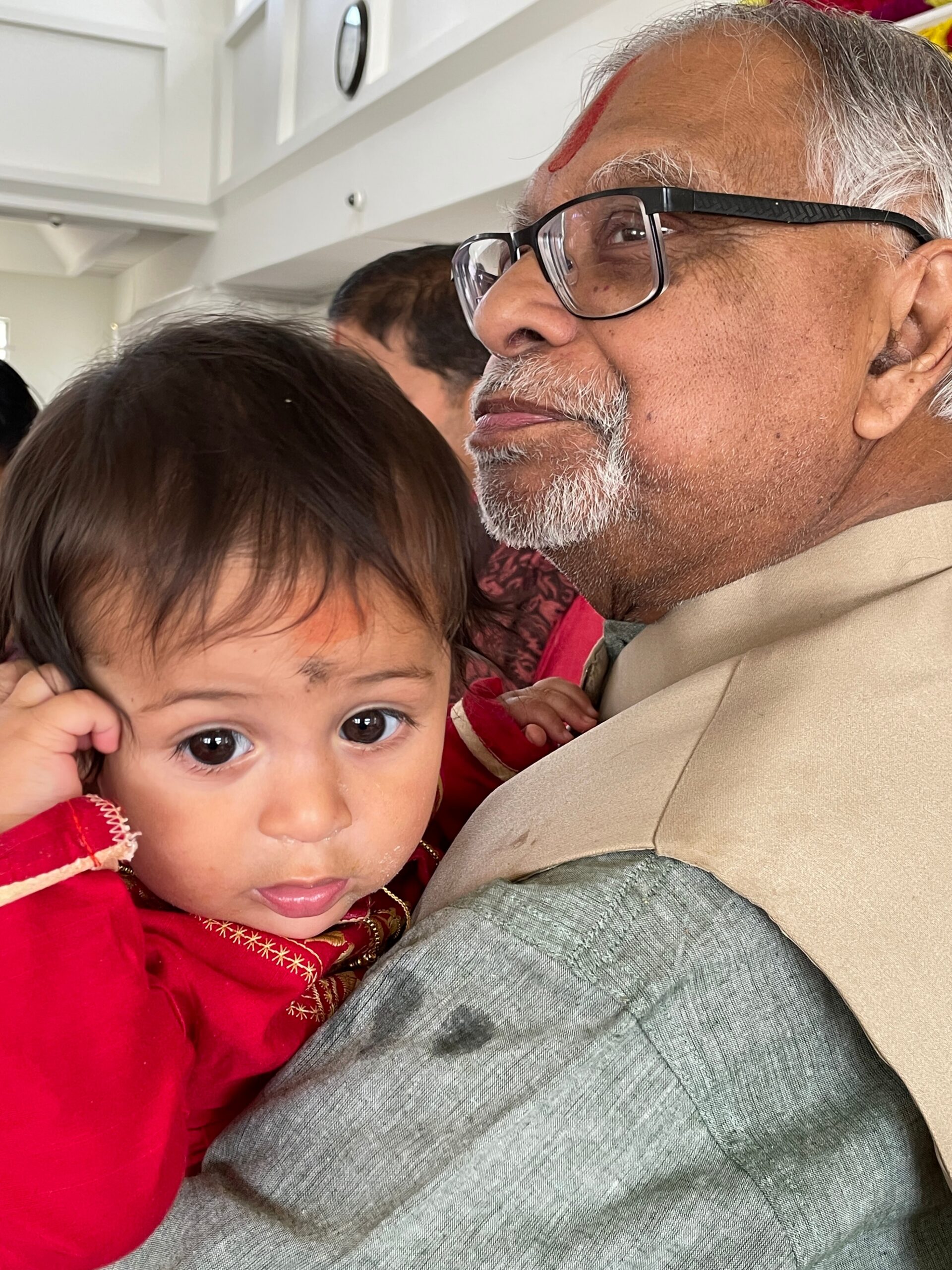 Close-up of an older man with glasses holding a young child in a red outfit; both face the camera indoors.