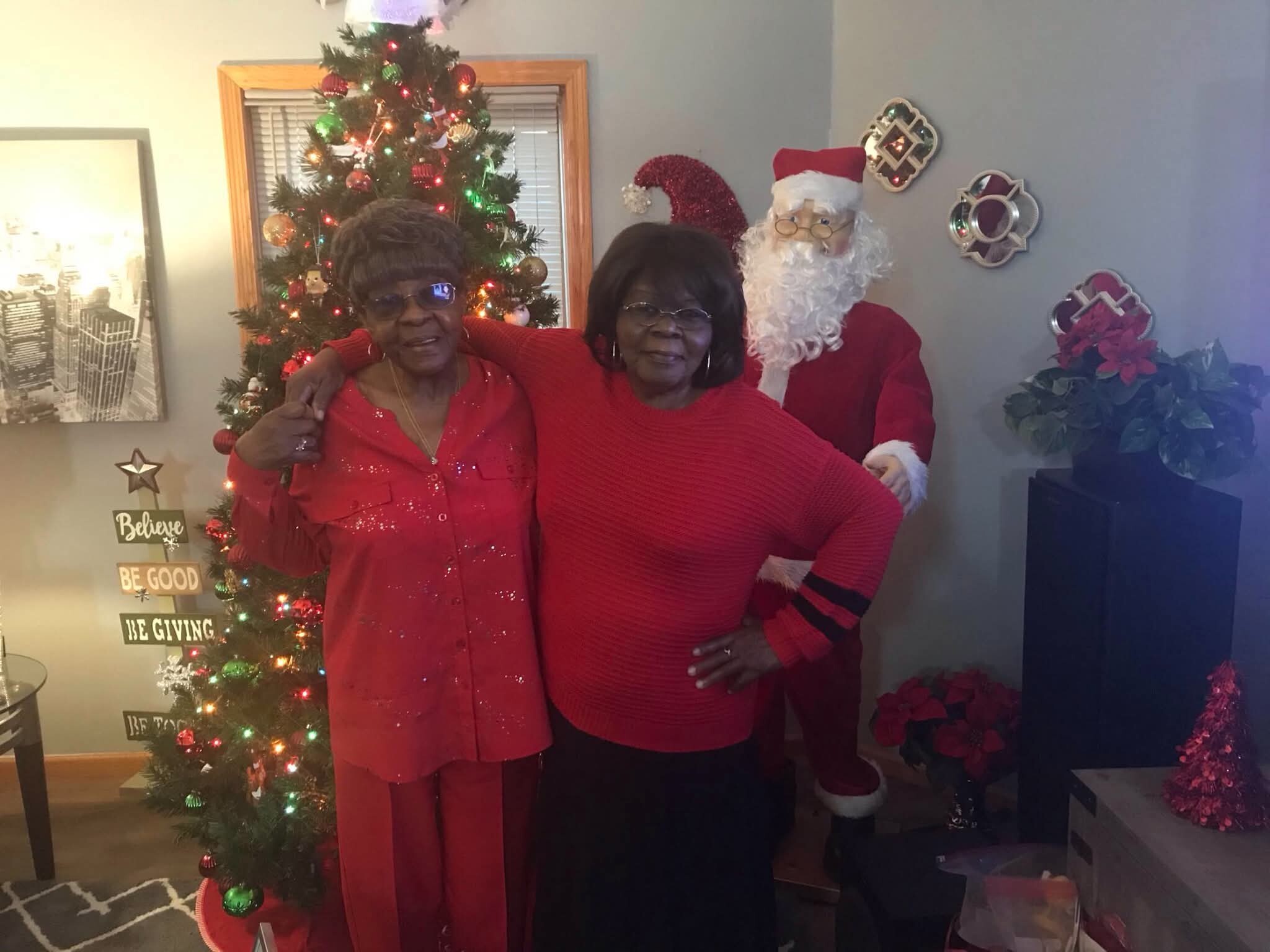 Two elderly women in red outfits pose with a Santa figure beside a lit Christmas tree in a living room.