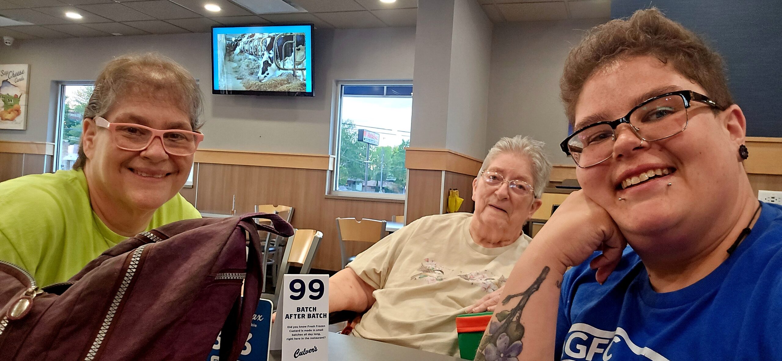 Three friends sit at a table in a casual restaurant, smiling at the camera; a '99' table sign is visible in front of them.