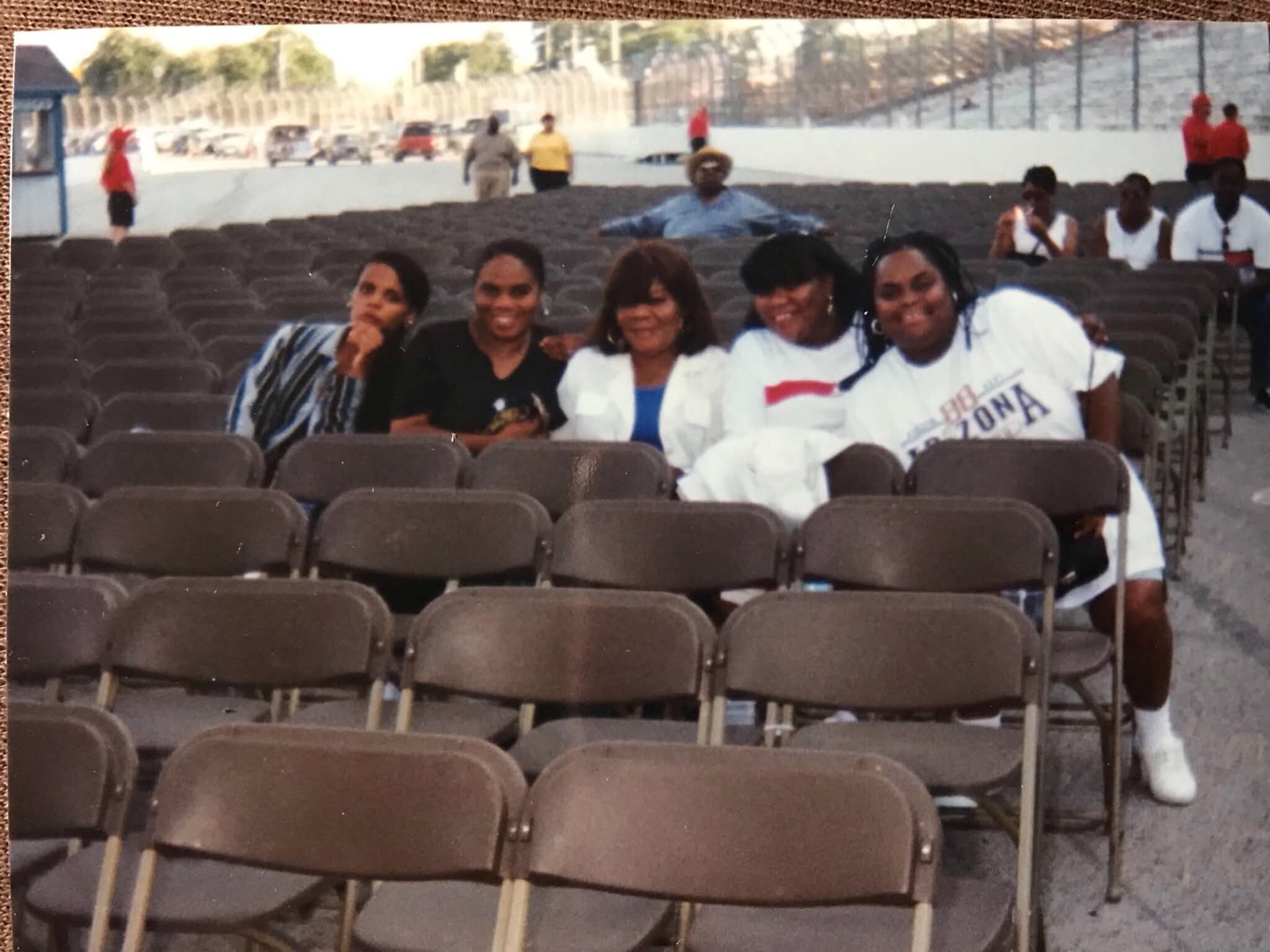 Four women sit in brown stadium seats, smiling at the camera; an outdoor arena with a track and people in the background.
