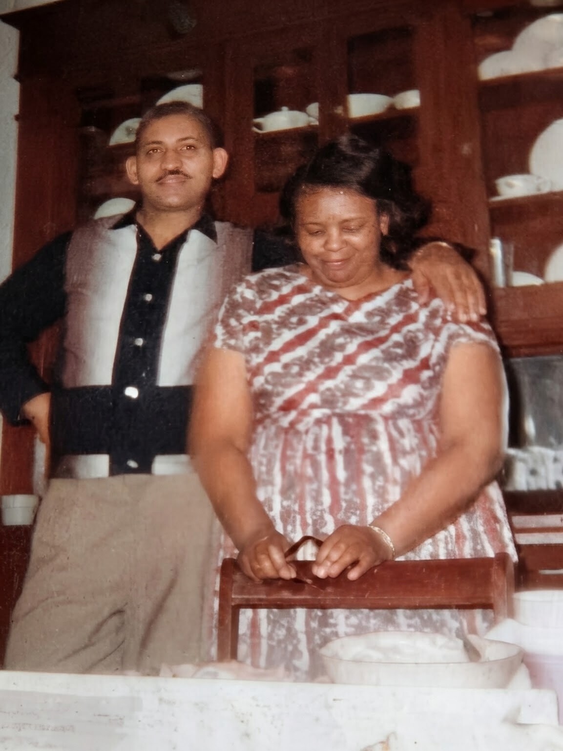Man with arm around a smiling woman in a kitchen/dining area, cabinets with dishes in the background, vintage photo feel.