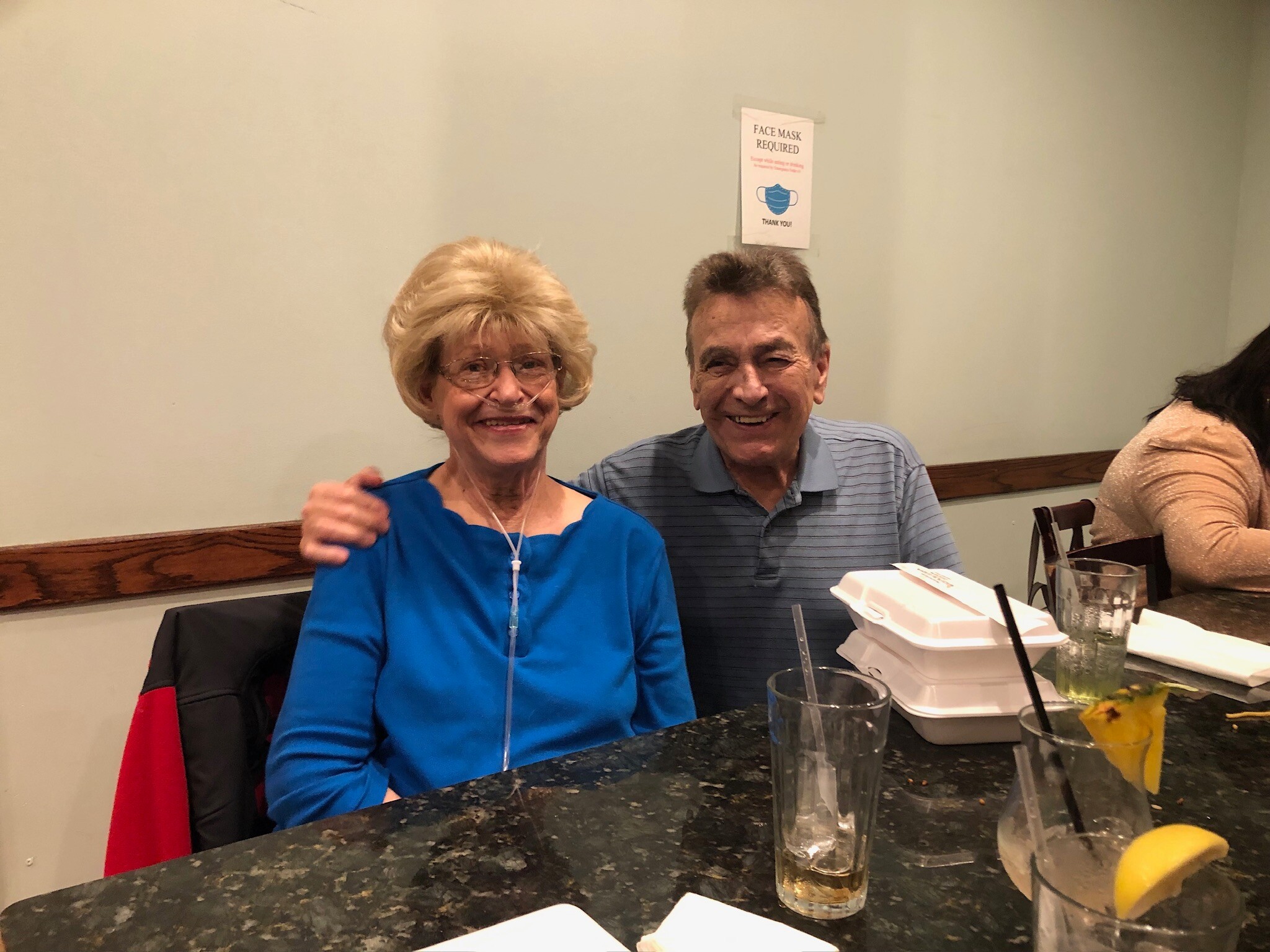 Smiling blonde woman in a blue blouse seated with a man in a striped polo at a restaurant table, drinks and a takeout box visible nearby.