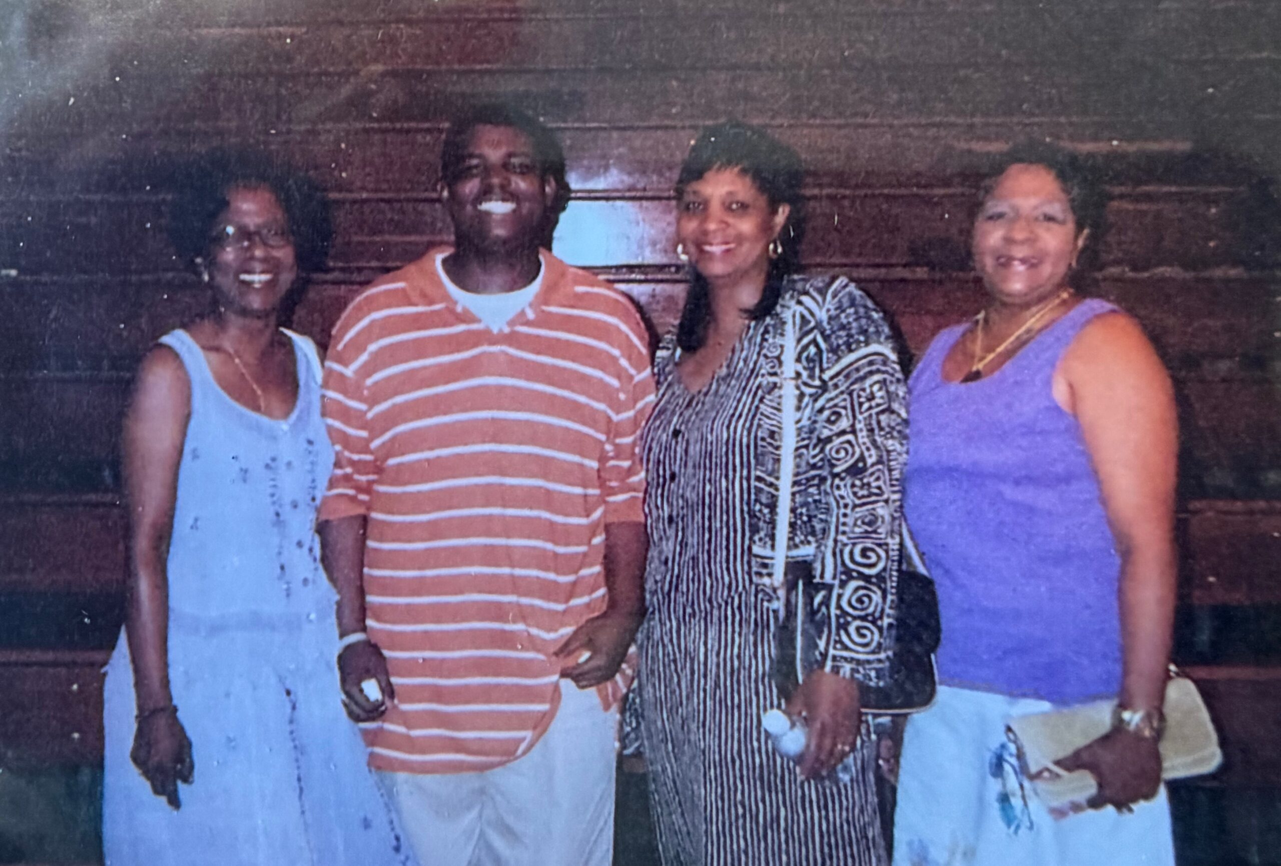 Four adults standing side by side and smiling for a family photo indoors, wearing colorful casual outfits.