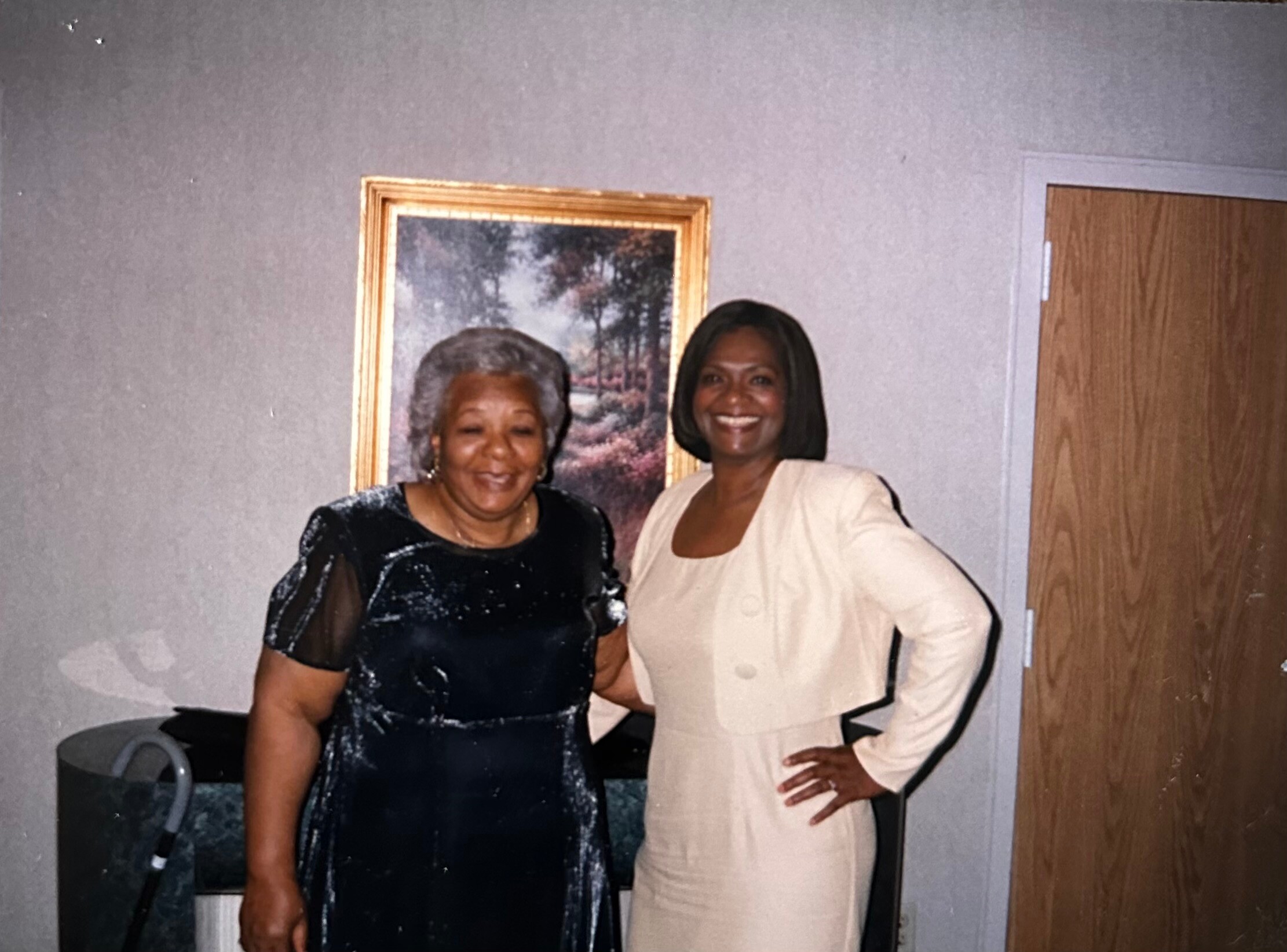 Two women in formal dresses posing indoors, smiling, beside a framed painting on a grey wall.