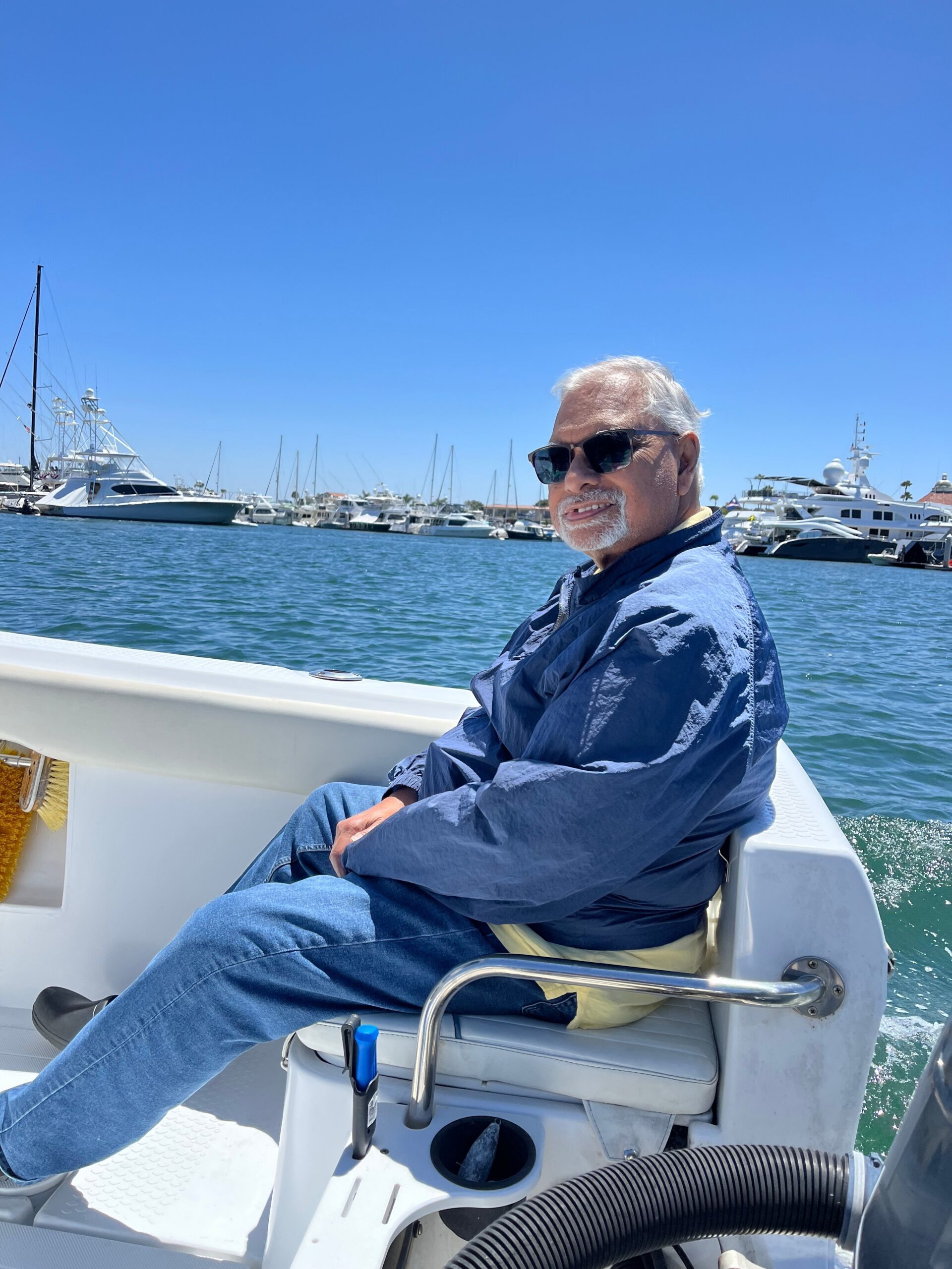 Man with gray hair and beard wearing sunglasses seated on a boat deck, marina and yachts in bright blue sky behind him.