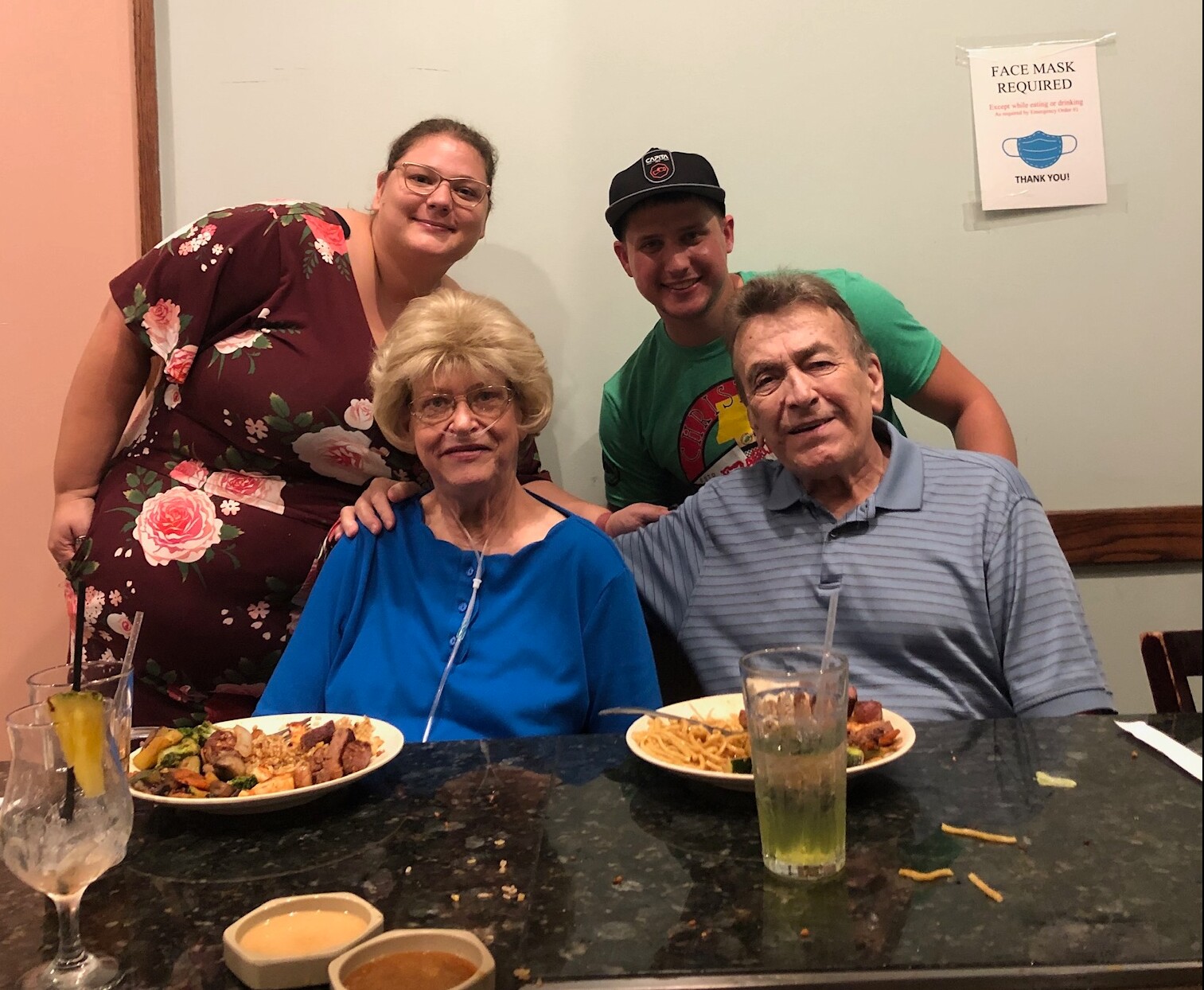 Four friends at a restaurant posing for a group photo, two seniors in front and two younger adults behind them, smiling with plates of food in front of them.