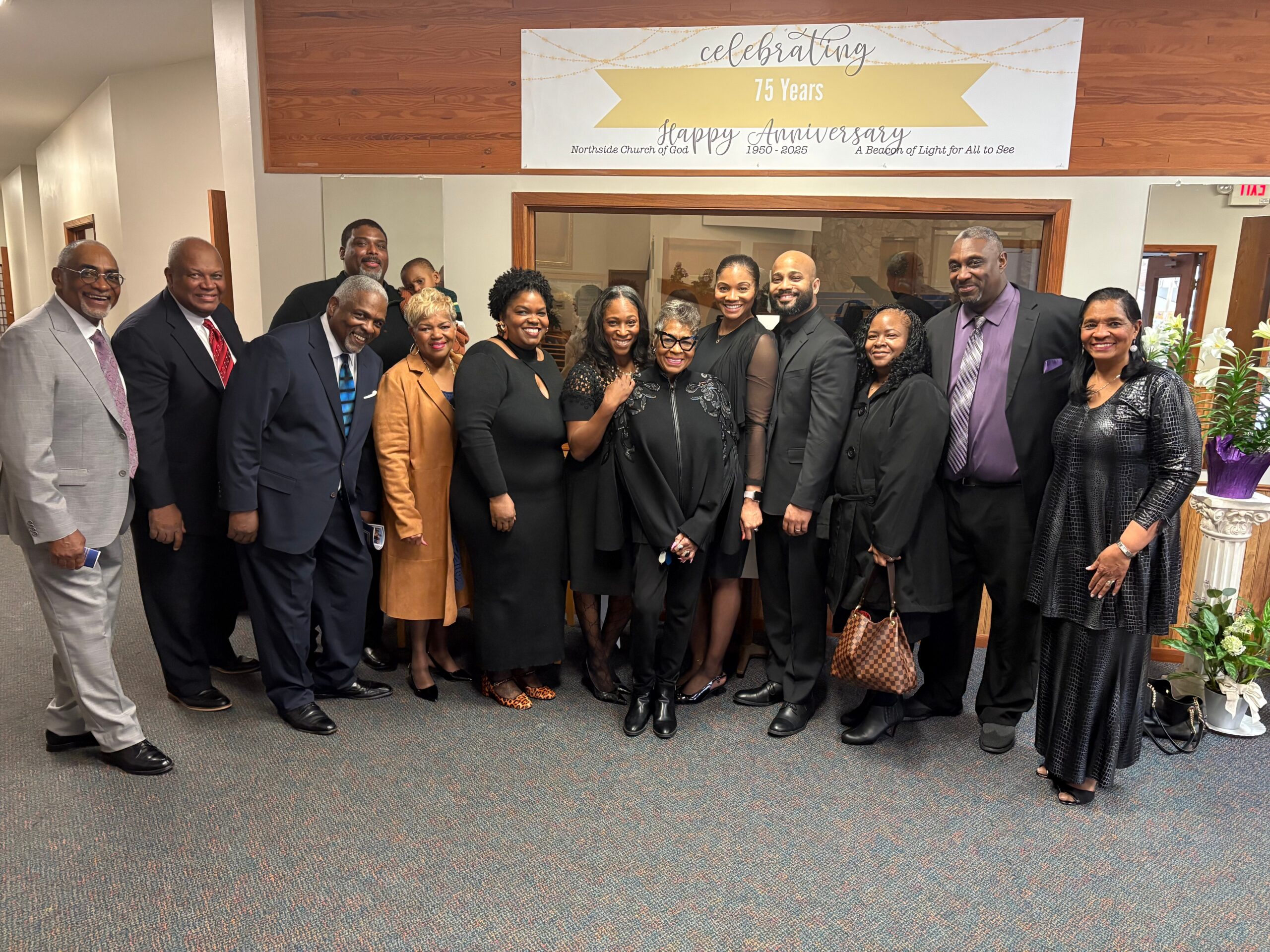 Group of adults in formal attire pose for a photo indoors under a '75 Years' anniversary banner at a church event.