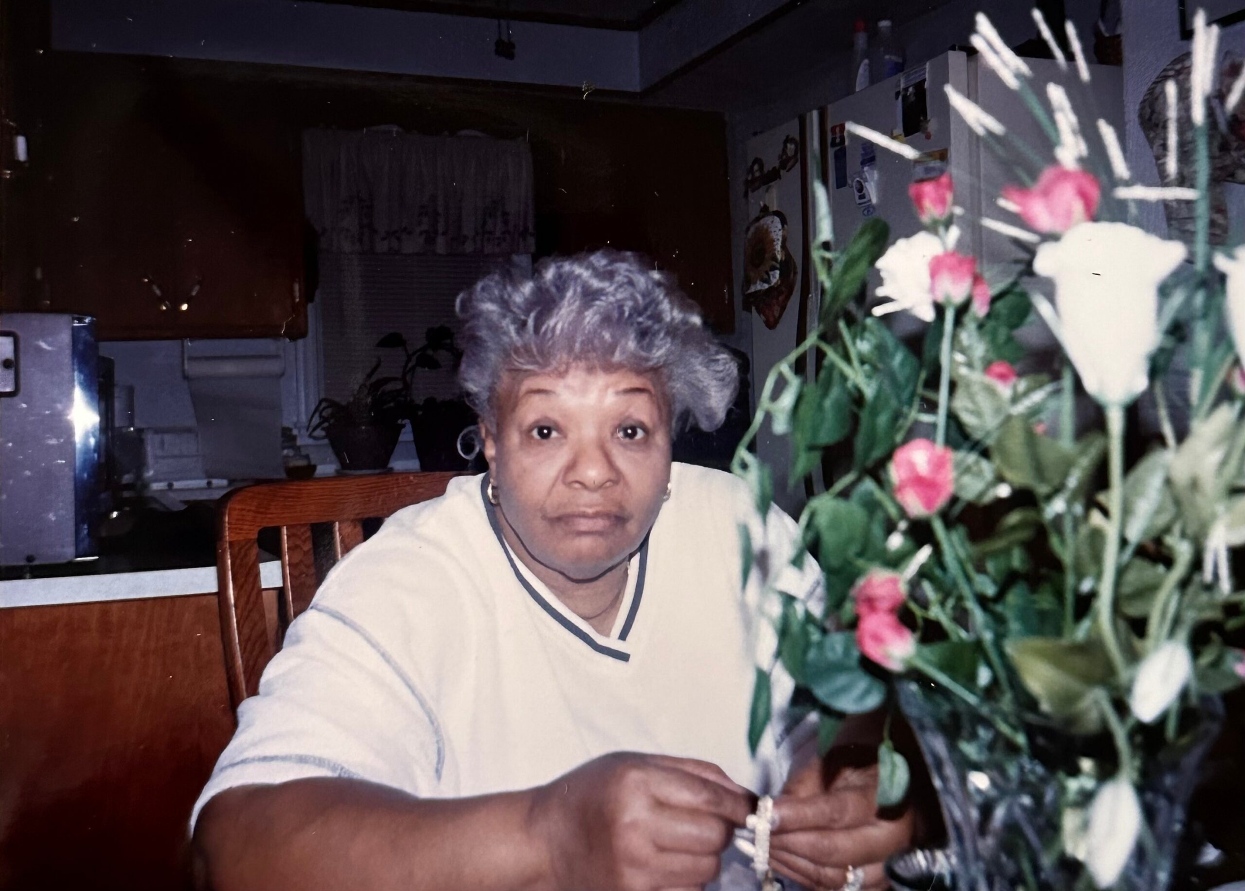 Older Black woman with short gray hair sits at a dining table, holding a rosary and looking toward the camera, with pink flowers in the foreground.