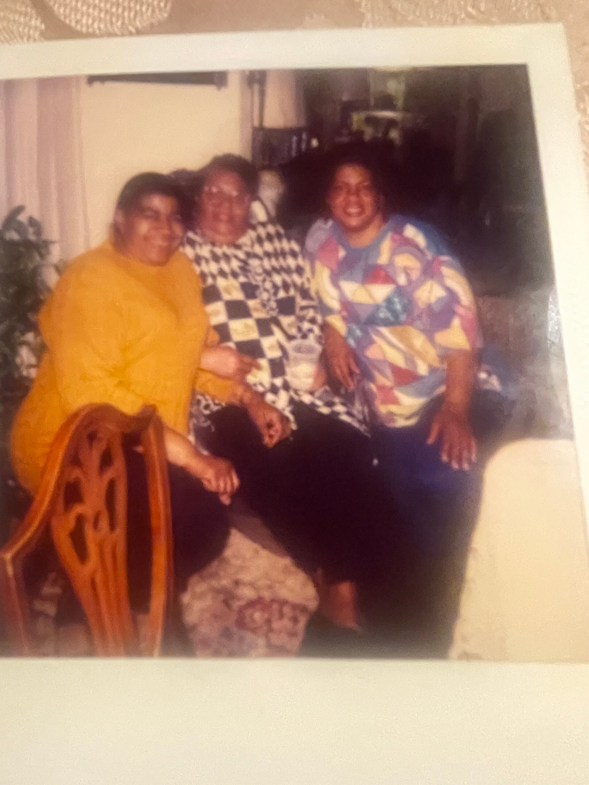 Three adults sit closely together on a couch indoors, smiling at the camera; left in a yellow top, middle in a black-and-white checkered shirt, right in a colorful geometric shirt.