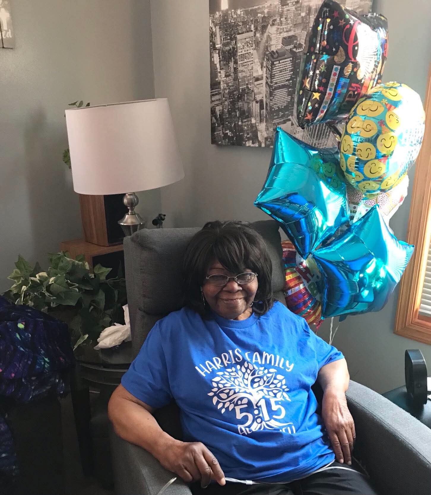 Smiling woman in a blue shirt sits in a chair at a celebration, with colorful balloons nearby in a living room.