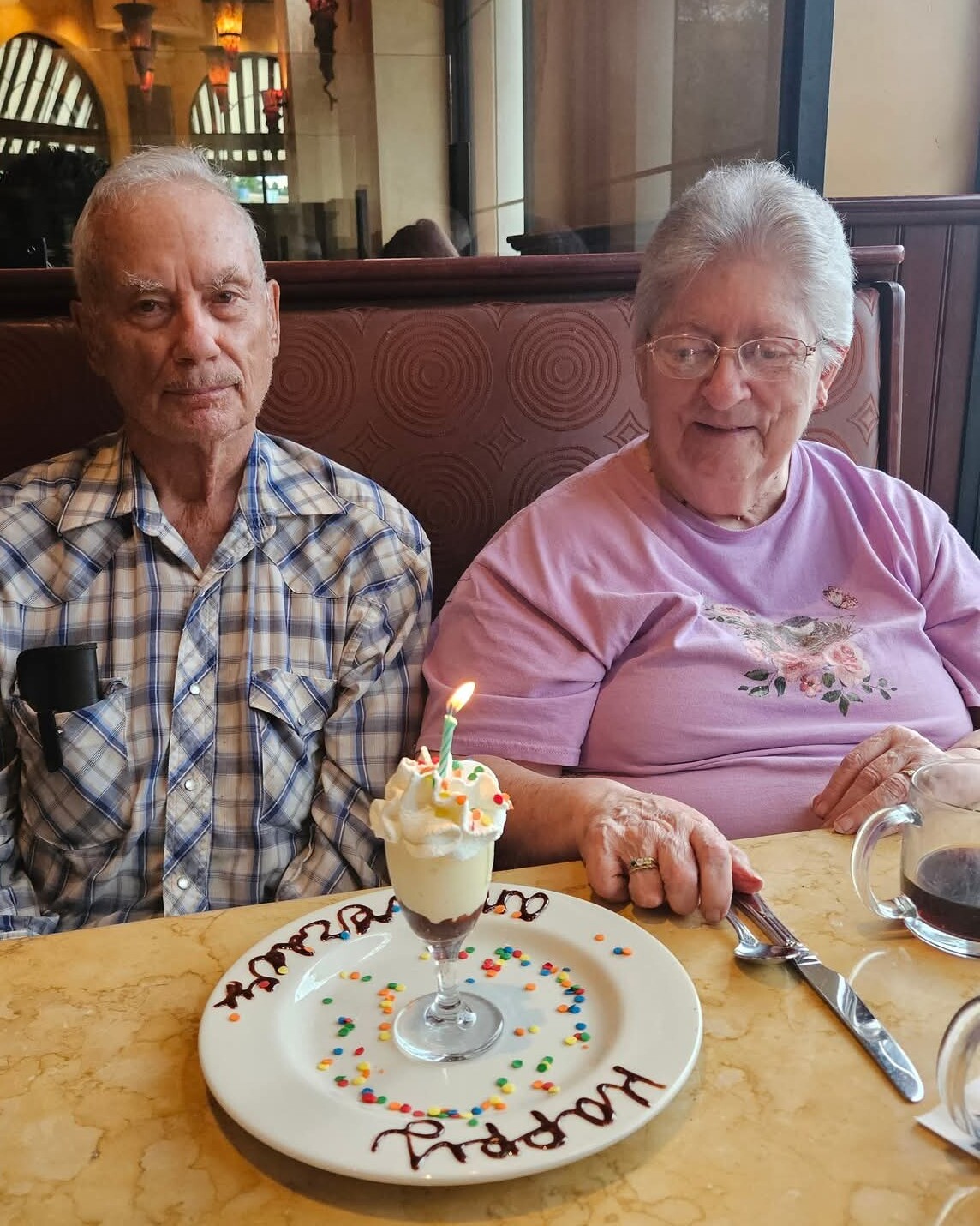 Elderly man and woman dining at a restaurant booth, with a birthday dessert and candle in front of them on the table.