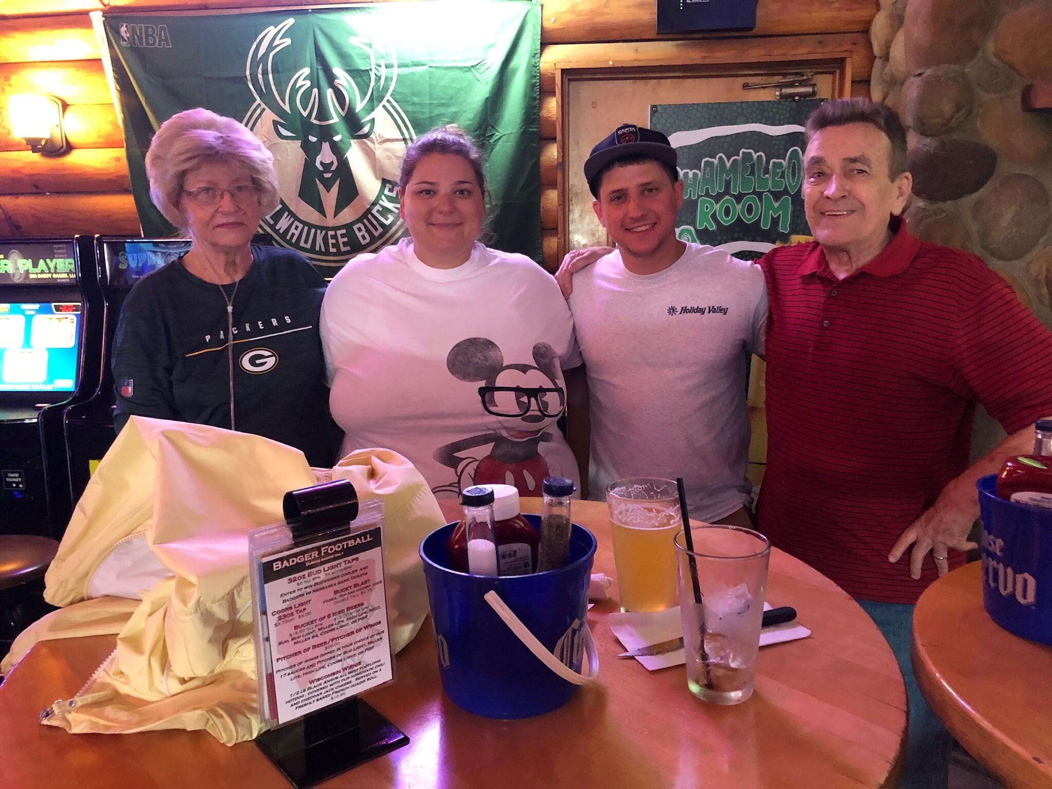 Four adults smiling and posing in a rustic bar, with a green Milwaukee Bucks banner in the background and drinks on the table.